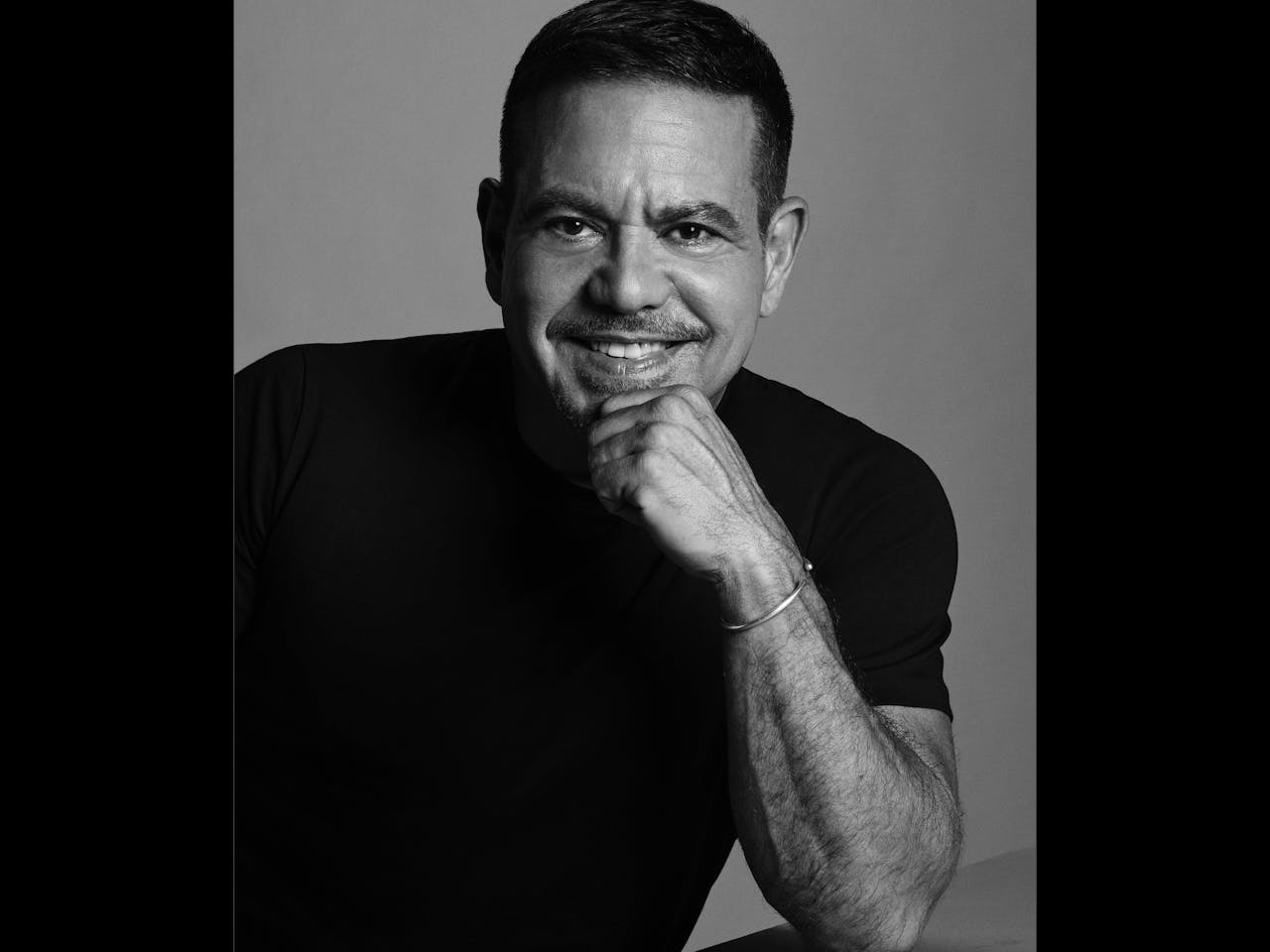 A black and white headshot of the designer and costume designer Narciso Rodriguez. He stands against a light backdrop, wears a black tshirt and holds his right hand up to his chin.