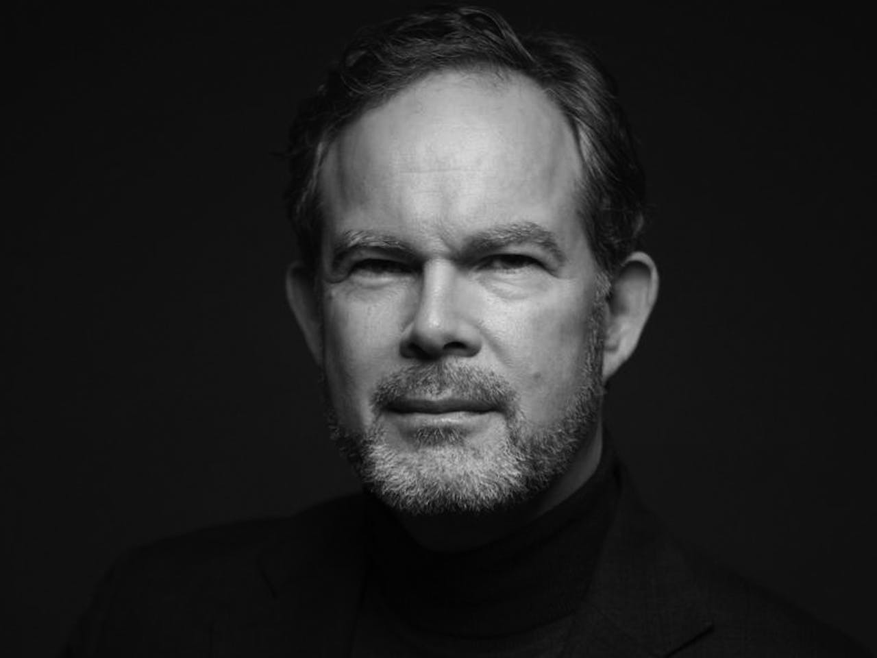 A black and white headshot of the opera singer Gerald Finley. He stands against a black backdrop wearing a black suit jacket and rollneck sweater.