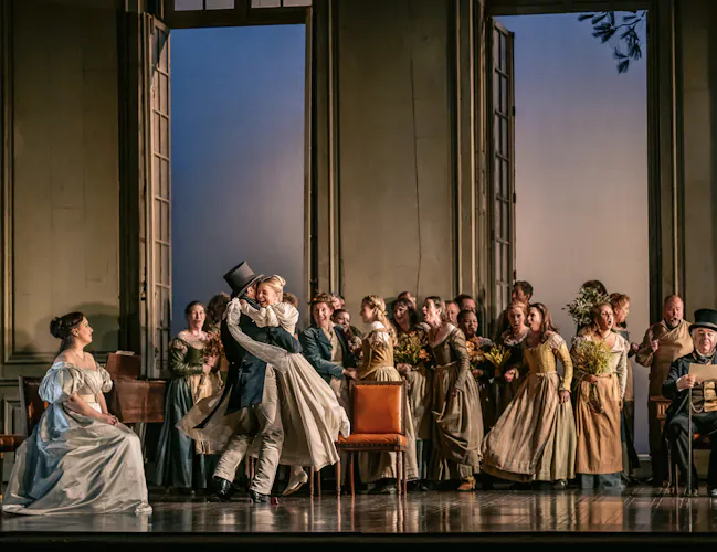 A group of people stand in front of large windows showing a sunset. A man in a blue suit and grey tophat has picked up and is spinning around a woman in a white dress.