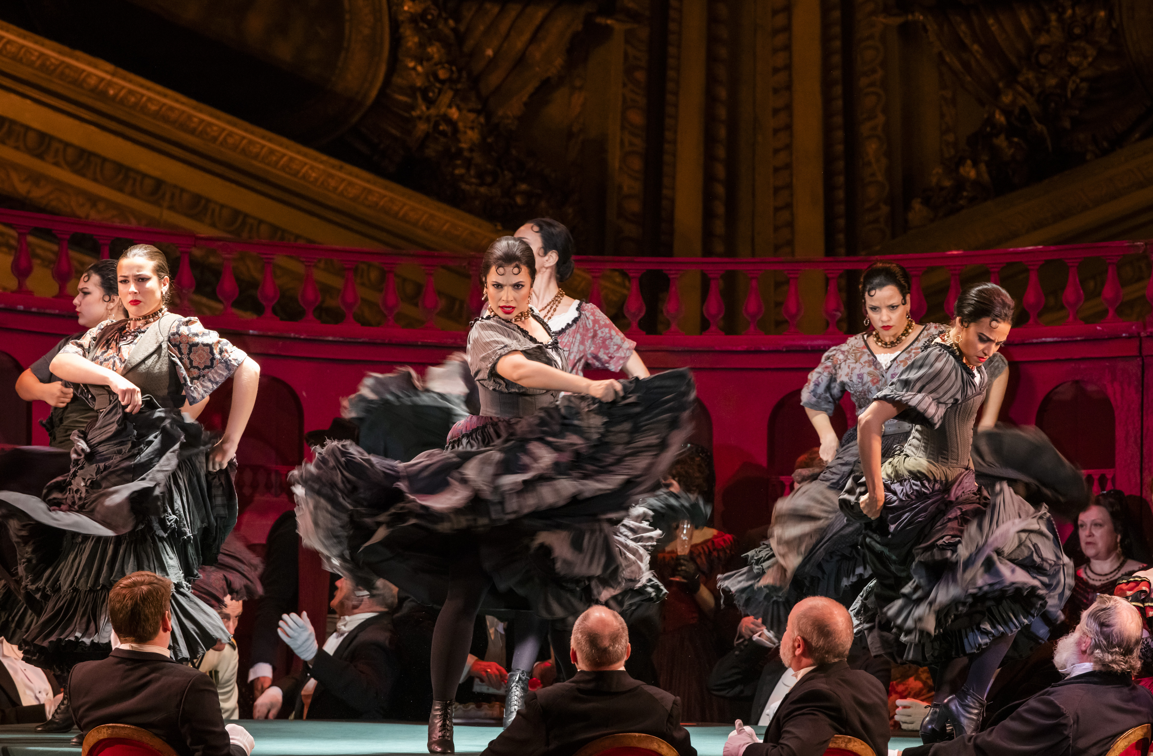 Three flamenco dancers in black dresses perform on stage with 3 male flamenco dancers behind them.