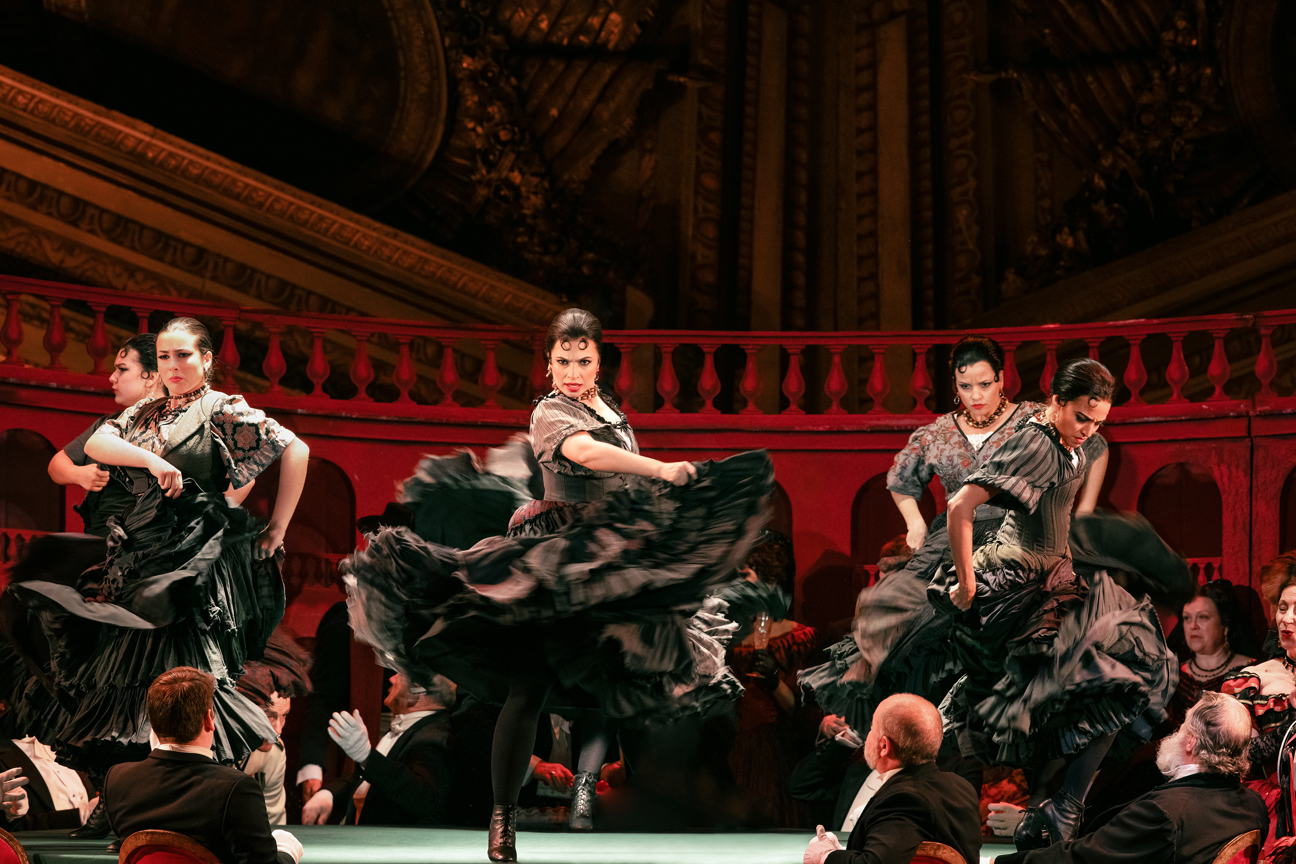 Flamenco dancers in black dresses swirl their skirts around them in a passionate dance. The wall behind them is red. 
