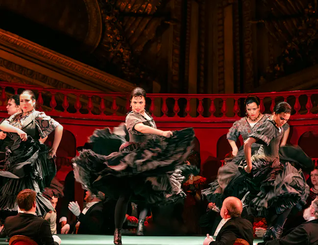 Flamenco dancers in black dresses swirl their skirts around them in a passionate dance. The wall behind them is red.