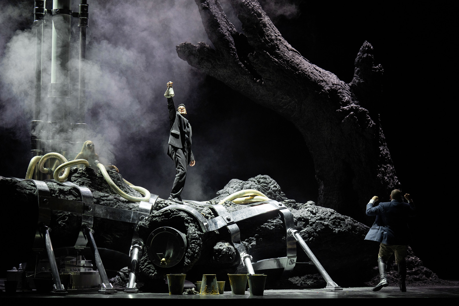 A character stands on a huge tree trunk in the centre of a darkly-lit stage, holding up a bag and singing. Another character stands at the root of the tree, looking up.