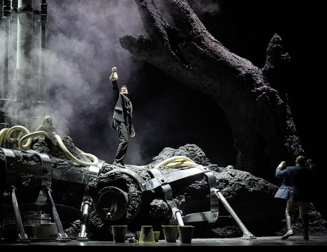 A character stands on a huge tree trunk in the centre of a darkly-lit stage, holding up a bag and singing. Another character stands at the root of the tree, looking up.