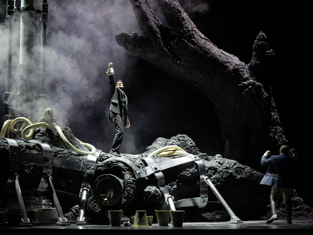 A character stands on a huge tree trunk in the centre of a darkly-lit stage, holding up a bag and singing. Another character stands at the root of the tree, looking up.