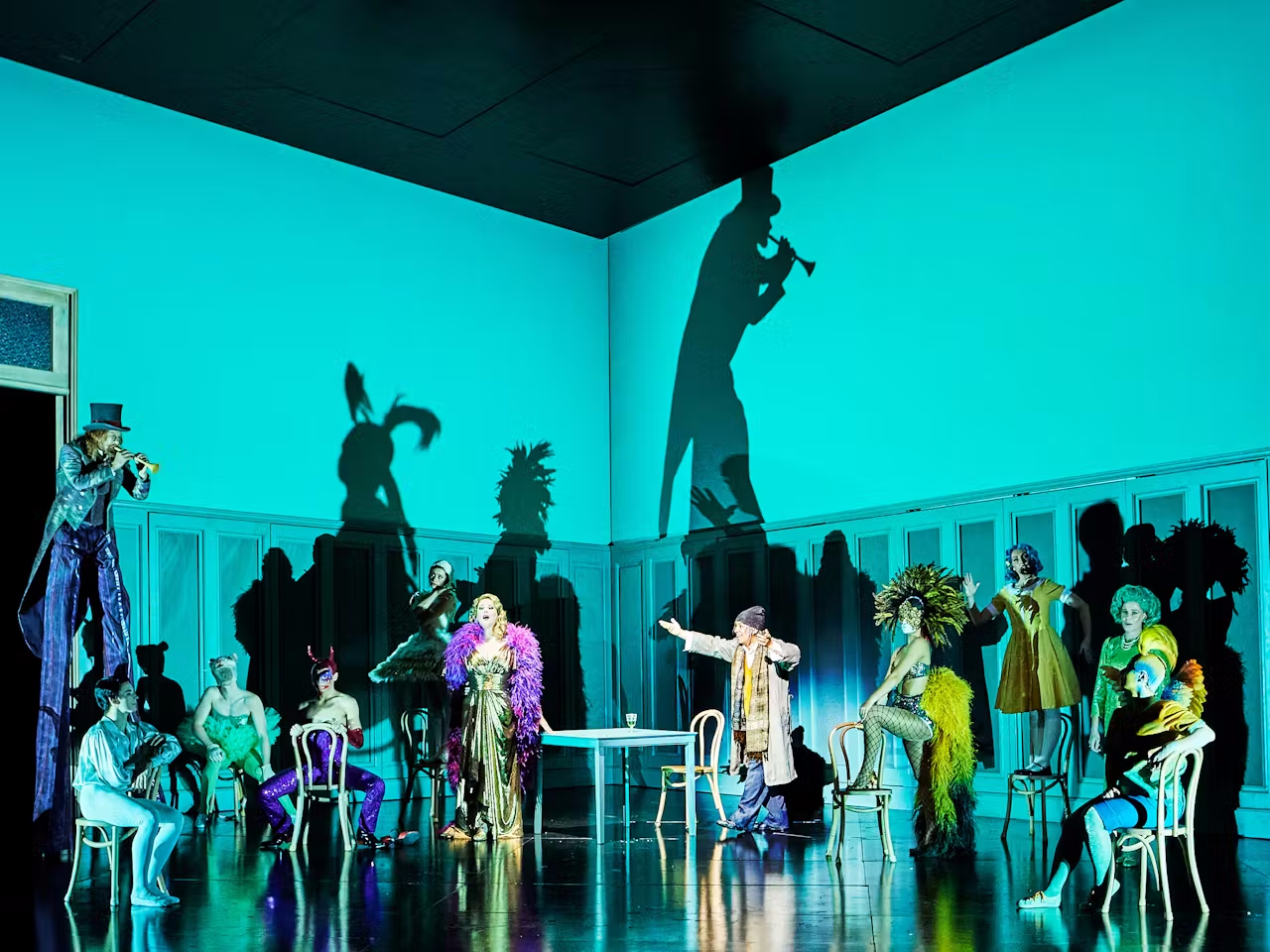 The stage is filled with a variety of characters in bright costumes and outfits - a singer wearing a gold dress and purple feather boa stands in the centre while people on stilts playing trumpets are lit creating shadows.