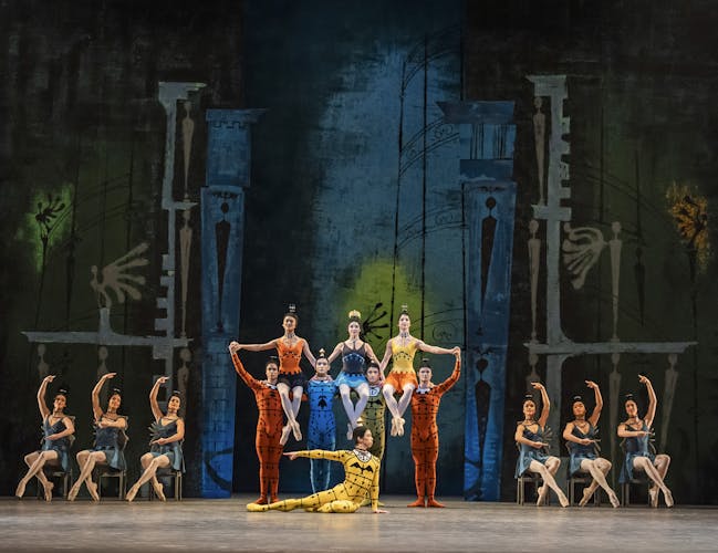 Dancers in bright costumes create formations; some seated, some sitting on the shoulders of others.