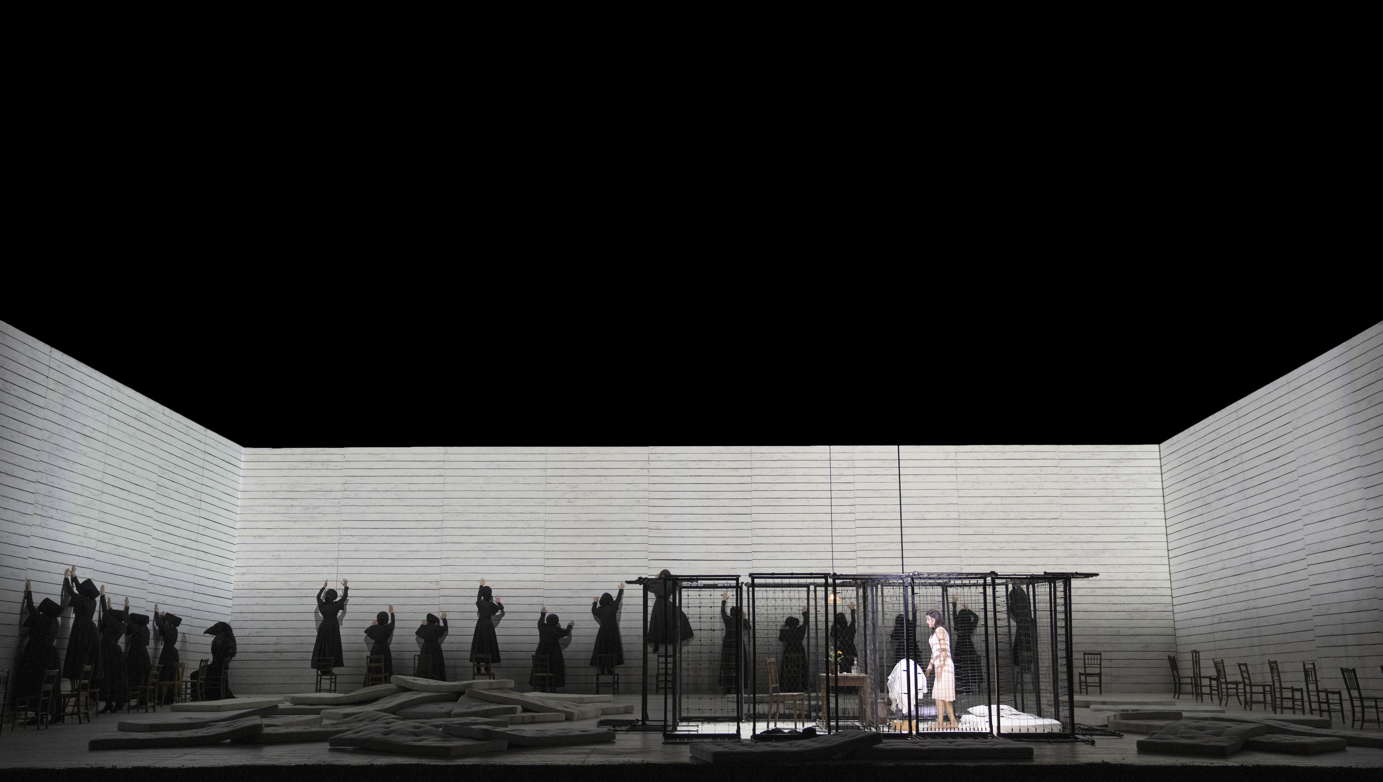 Several actors in black cloaks put their arms up to three white walls with their back to the audience. There is a cage with a woman in a white dress in the centre of the stage.