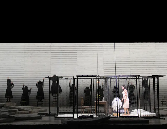 Several actors in black cloaks put their arms up to three white walls with their back to the audience. There is a cage with a woman in a white dress in the centre of the stage.