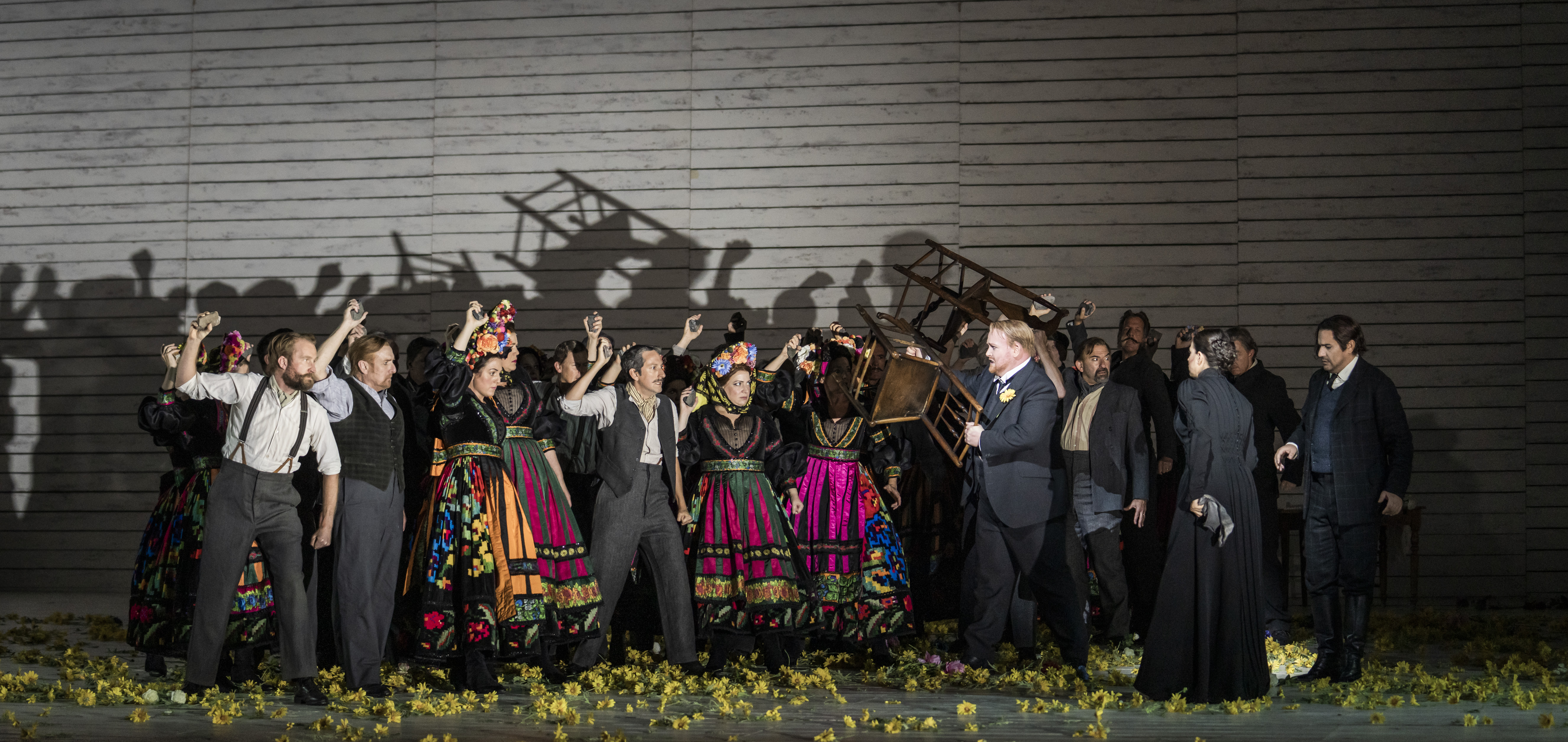 Lots of people stand on stage either holding up chairs or their fists. Four of them are women in brightly coloured traditional clothing.