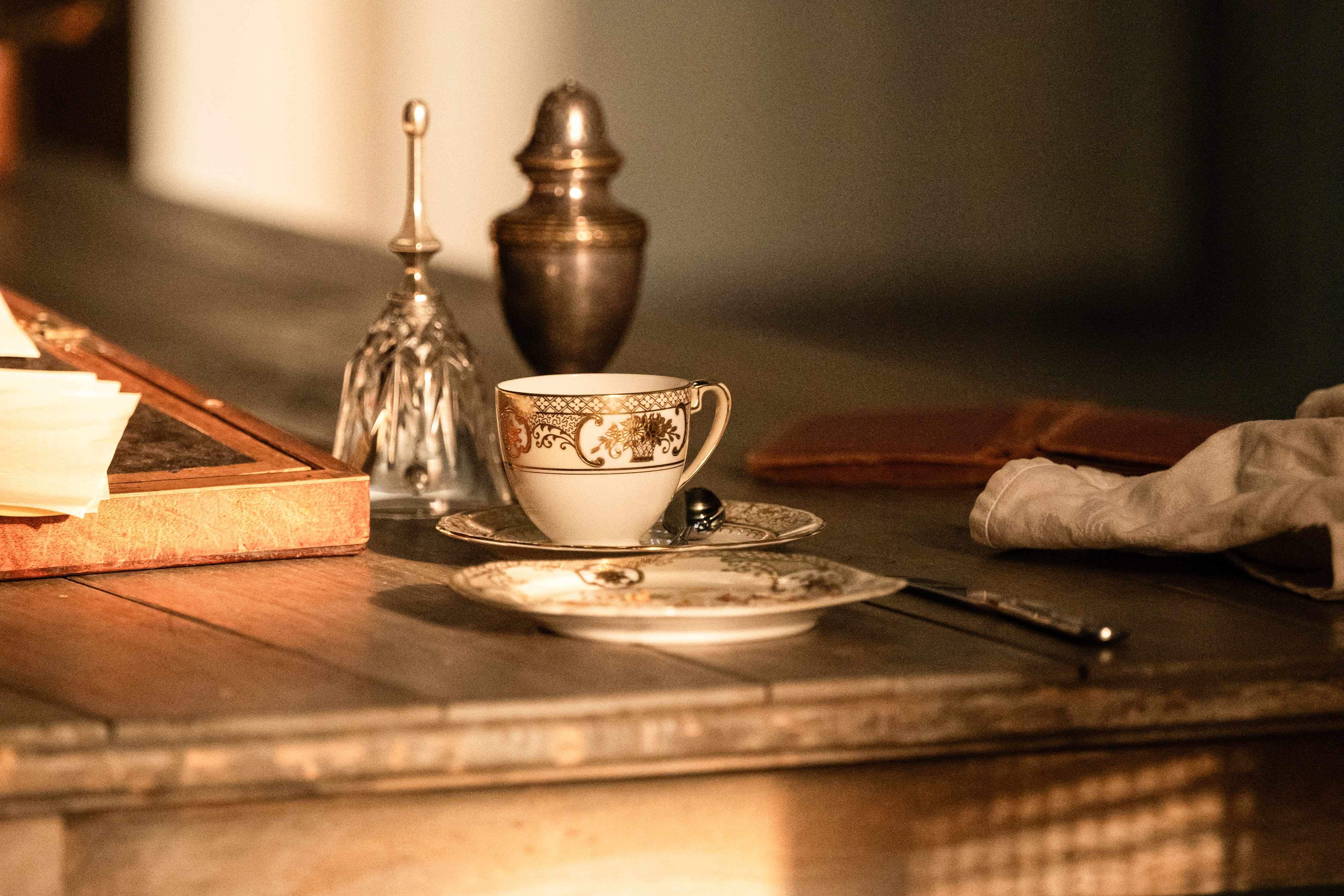 A wooden table in sunlight set with a china teacup and saucer and a spoon placed alongside, a china plate, a knife, a glass bell, a brass urn and a white glove.