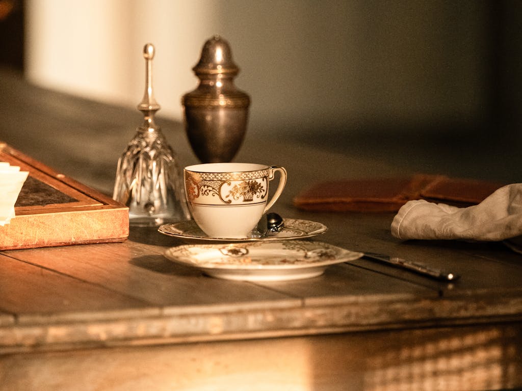 A wooden table in sunlight set with a china teacup and saucer and a spoon placed alongside, a china plate, a knife, a glass bell, a brass urn and a white glove.