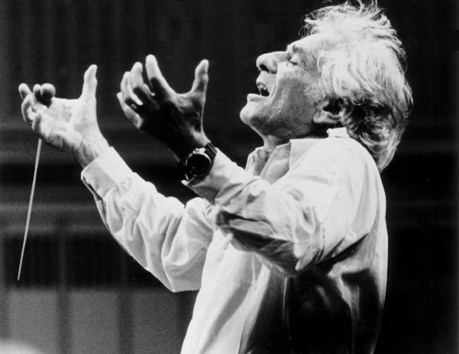 Leonard Bernstein conducts an orchestra. Both his arms are raised and he is holding a baton in his right hand.