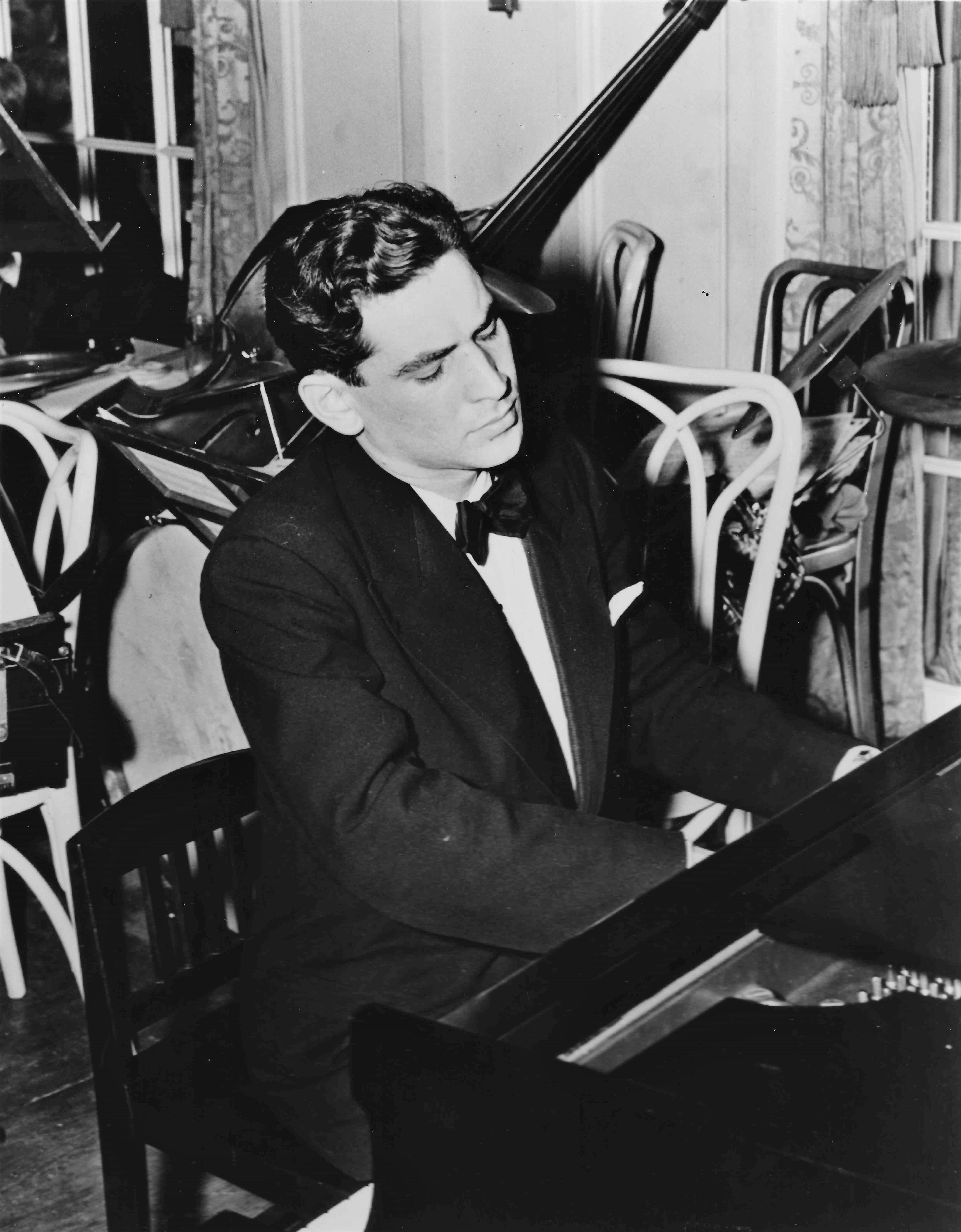 Leonard Bernstein is playing a piano. He is wearing a dinner suit and there are chairs stacked up in the background.