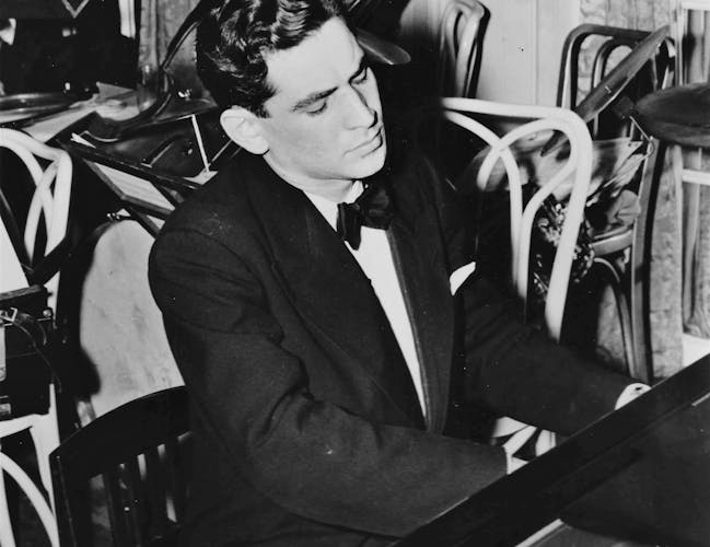 Leonard Bernstein is playing a piano. He is wearing a dinner suit and there are chairs stacked up in the background.