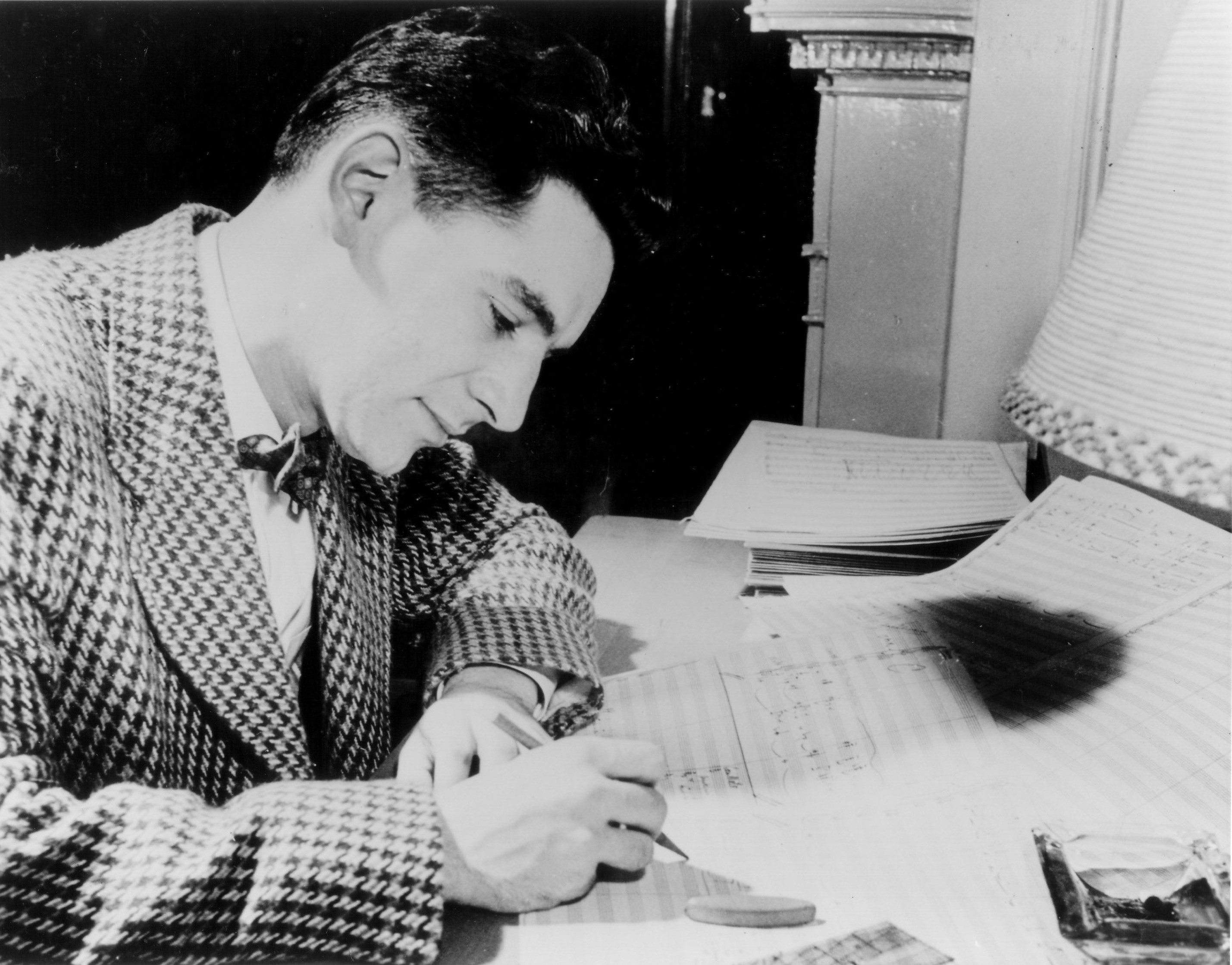 Leonard Bernstein, wearing a check blazer and bowtie, writes on a musical score.