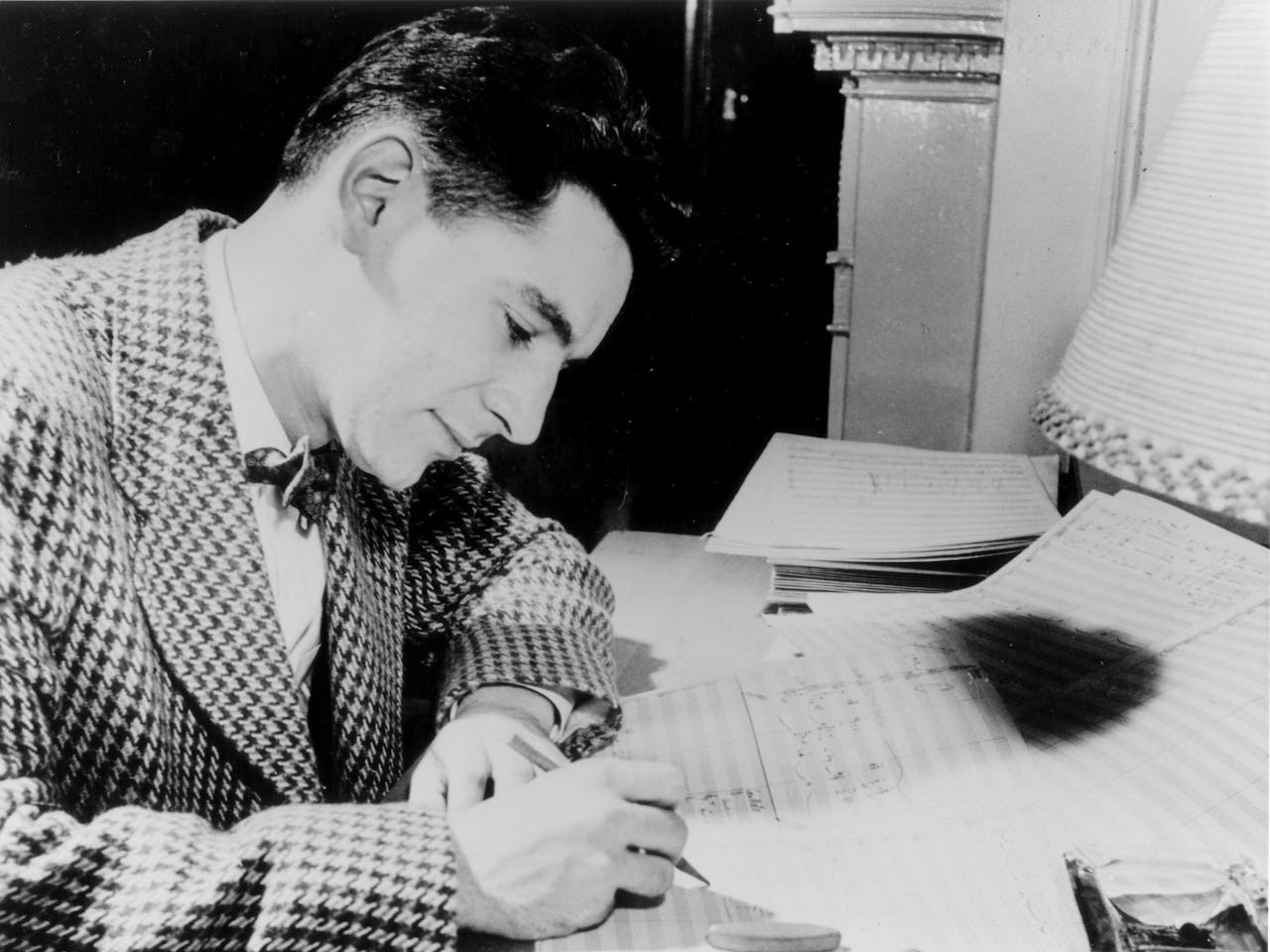 Leonard Bernstein, wearing a check blazer and bowtie, writes on a musical score.