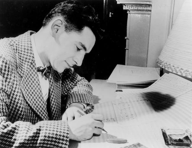 Leonard Bernstein, wearing a check blazer and bowtie, writes on a musical score.