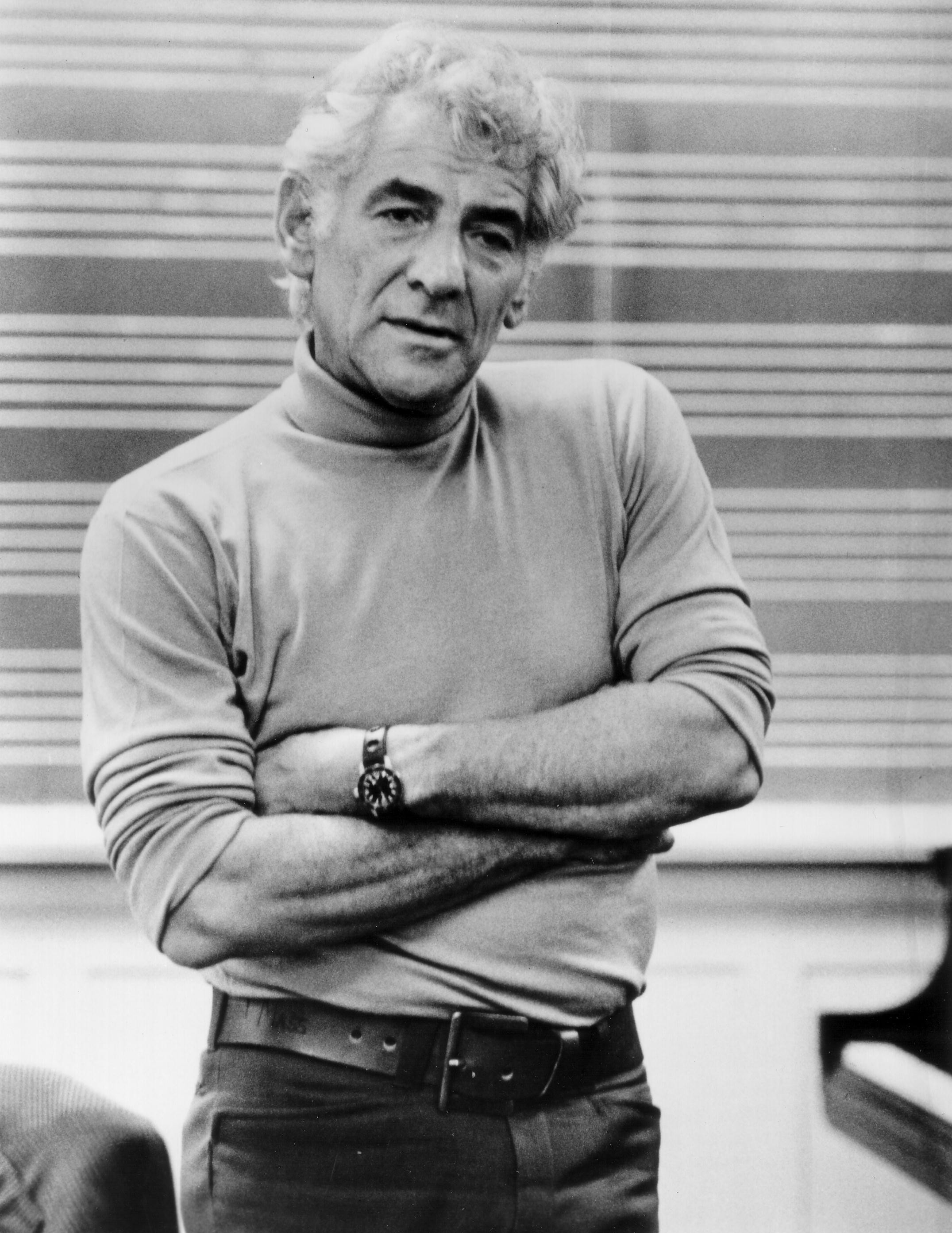 Leonard Bernstein stands facing the camera with a neutral expression. He is wearing a light turtle neck and jeans. His arms are folded.
