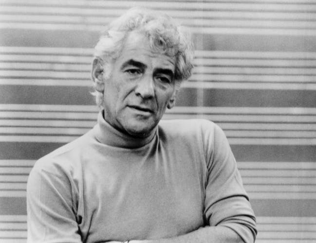 Leonard Bernstein stands facing the camera with a neutral expression. He is wearing a light turtle neck and jeans. His arms are folded.