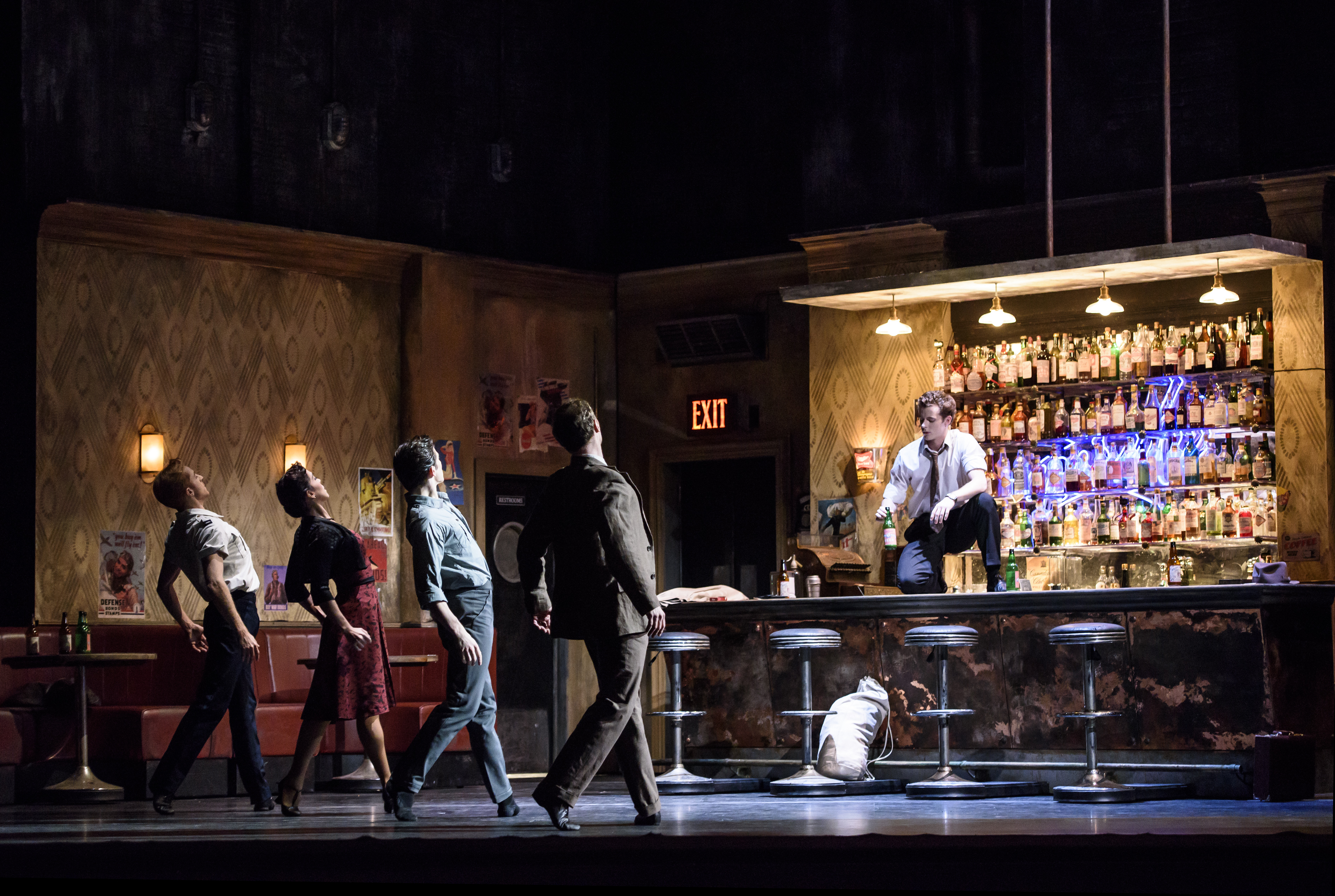 Four dancers stand facing a bar. Another male dancer sits on top of the bar, facing them.