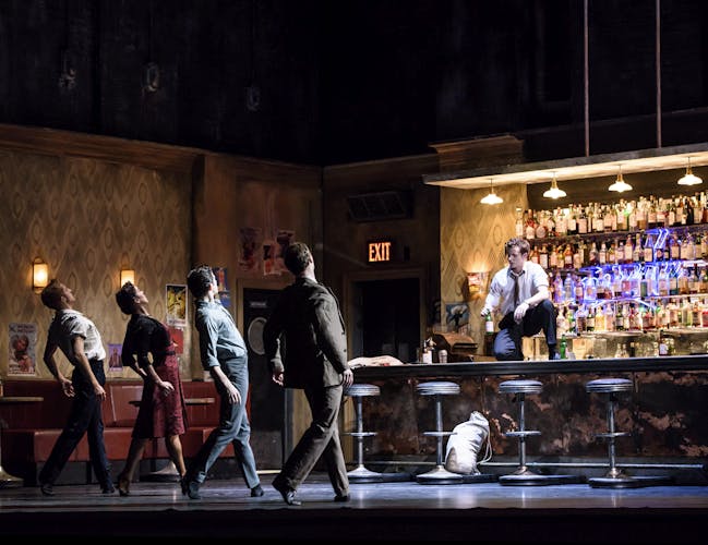 Four dancers stand facing a bar. Another male dancer sits on top of the bar, facing them.