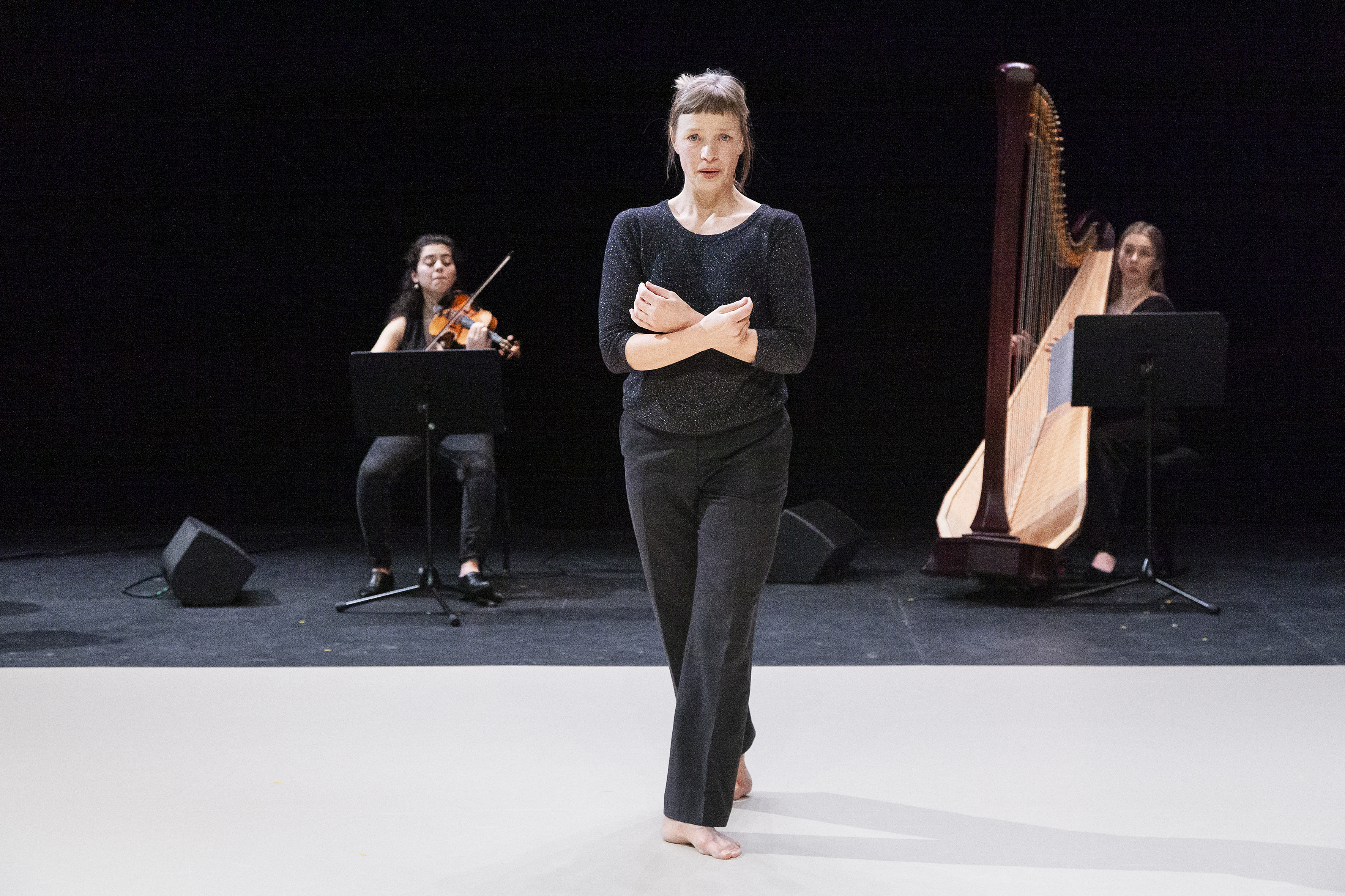 A dancer in black clothing stands on the stage at the fore of the photo. A violinist and a harpist are sat behind the dancer accompanying her.