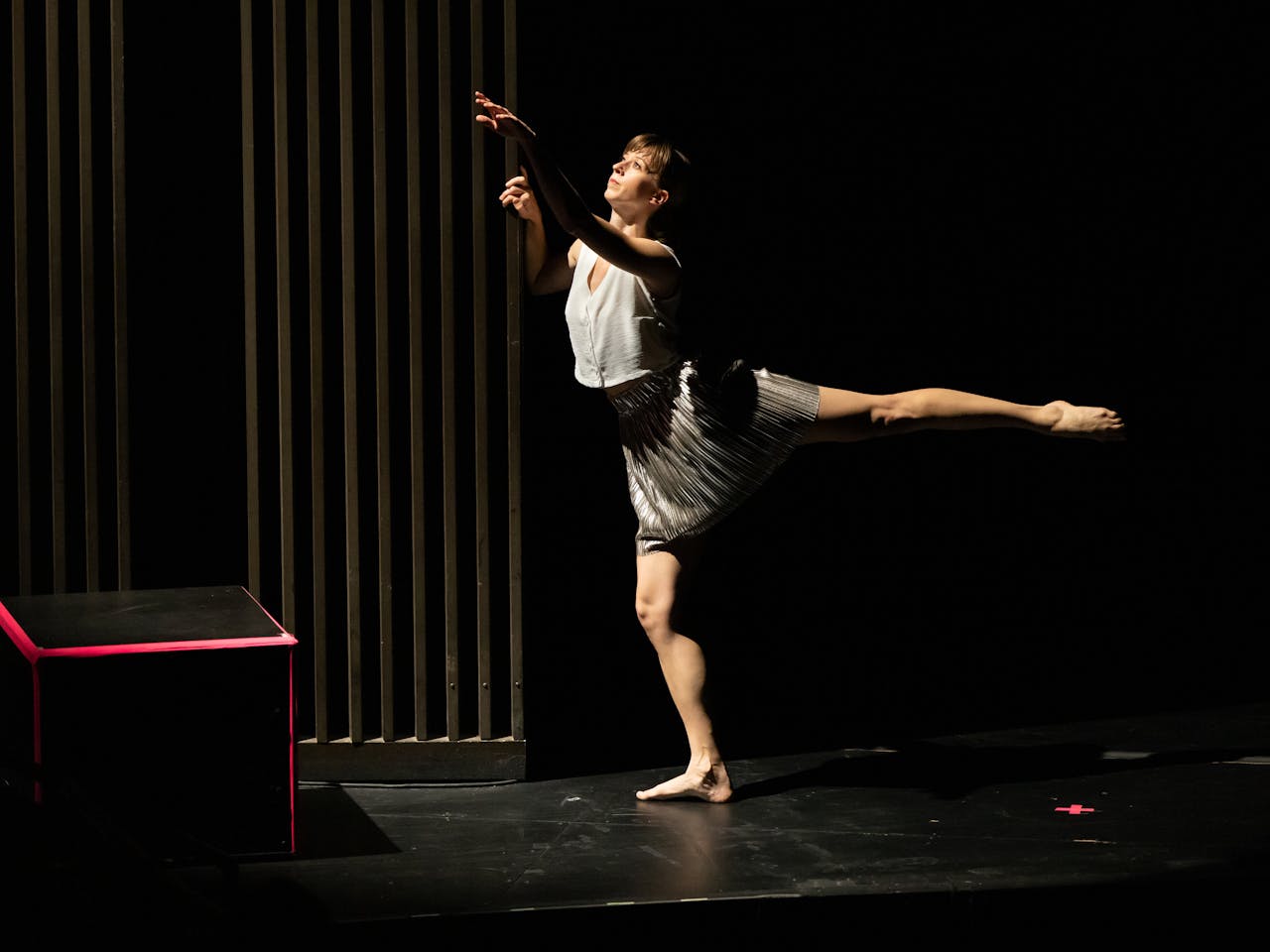A barefoot dancer, dressed in a grey button-up vest and skirt holds a position on a dark stage whilst standing on one leg.