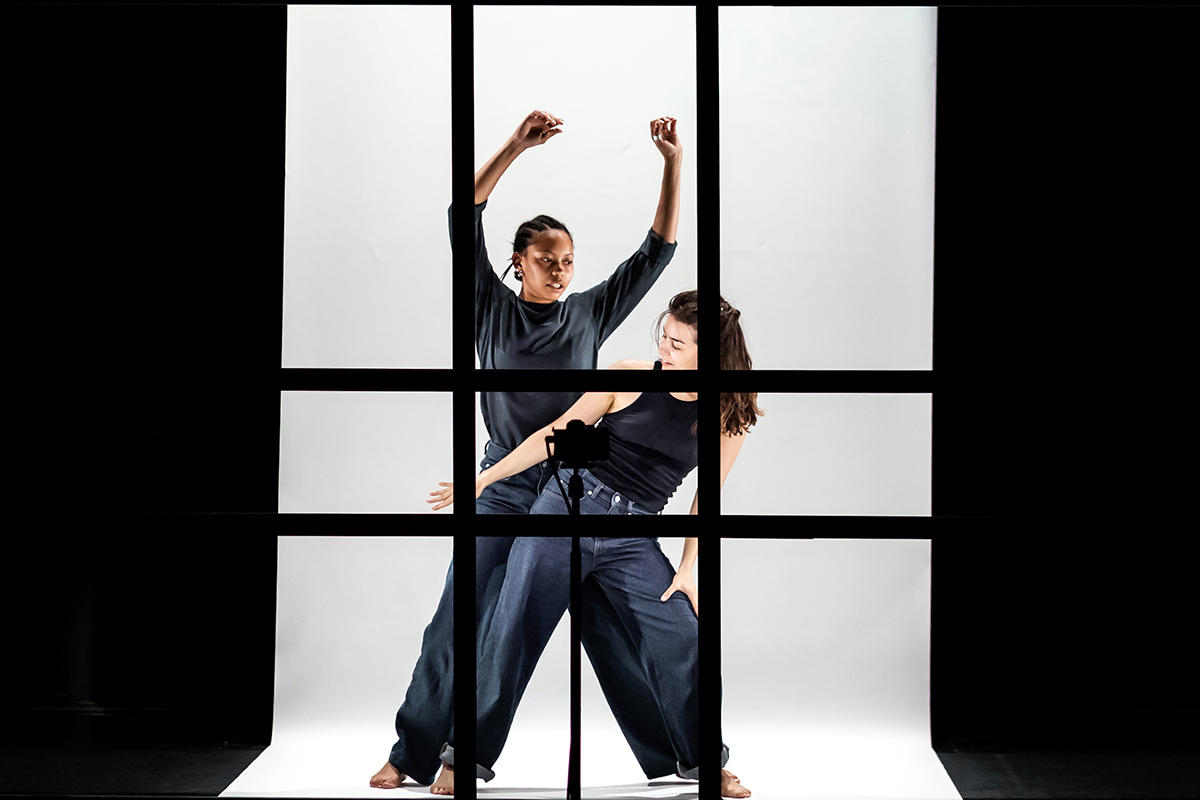 Two dancers dressed in black appear behind a series of black frames on a white background. One dancer is in front of the other. The dancer behind has their arms raised whilst the dancer in front leans to the side