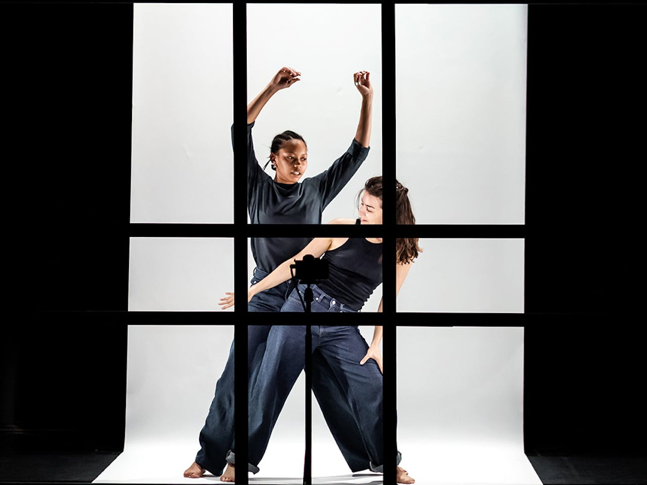 Two dancers dressed in black appear behind a series of black frames on a white background. One dancer is in front of the other. The dancer behind has their arms raised whilst the dancer in front leans to the side