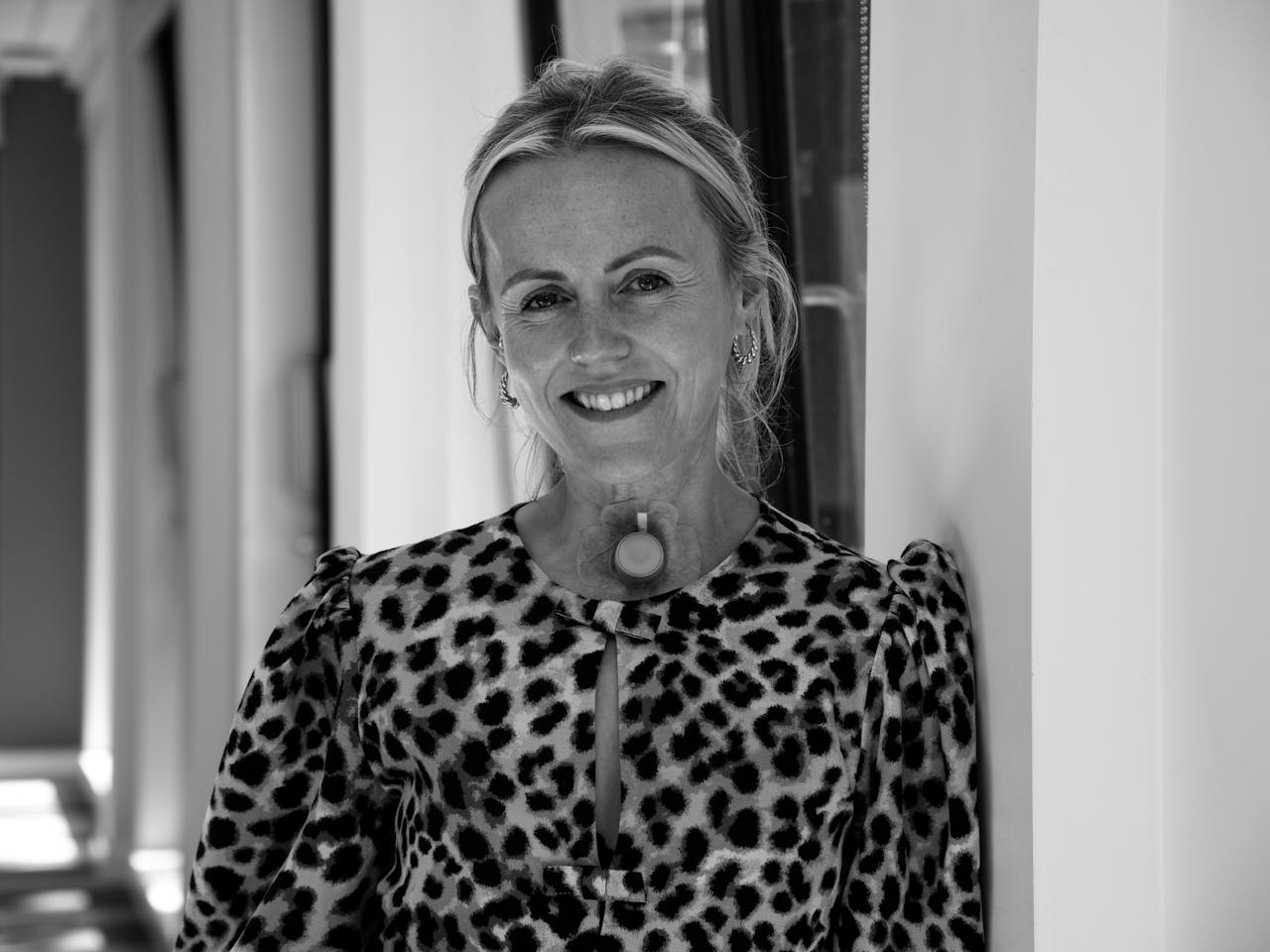 A black and white headshot of the cast member of Sound Voice Project Tanja Bage. She stands against a door in a brightly lit hallways wearing a leopard print top.