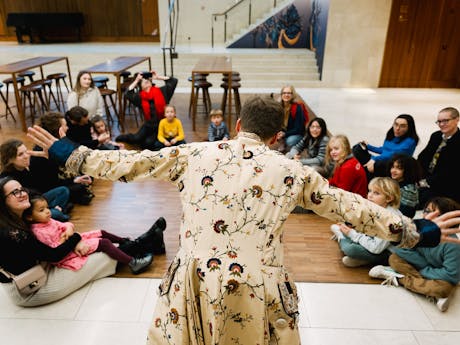 A man in a patterned coat stands with arms outstretched, addressing a seated group of adults and children in an open indoor space. The group appears engaged, sitting on a wooden floor with tables and chairs in the background.