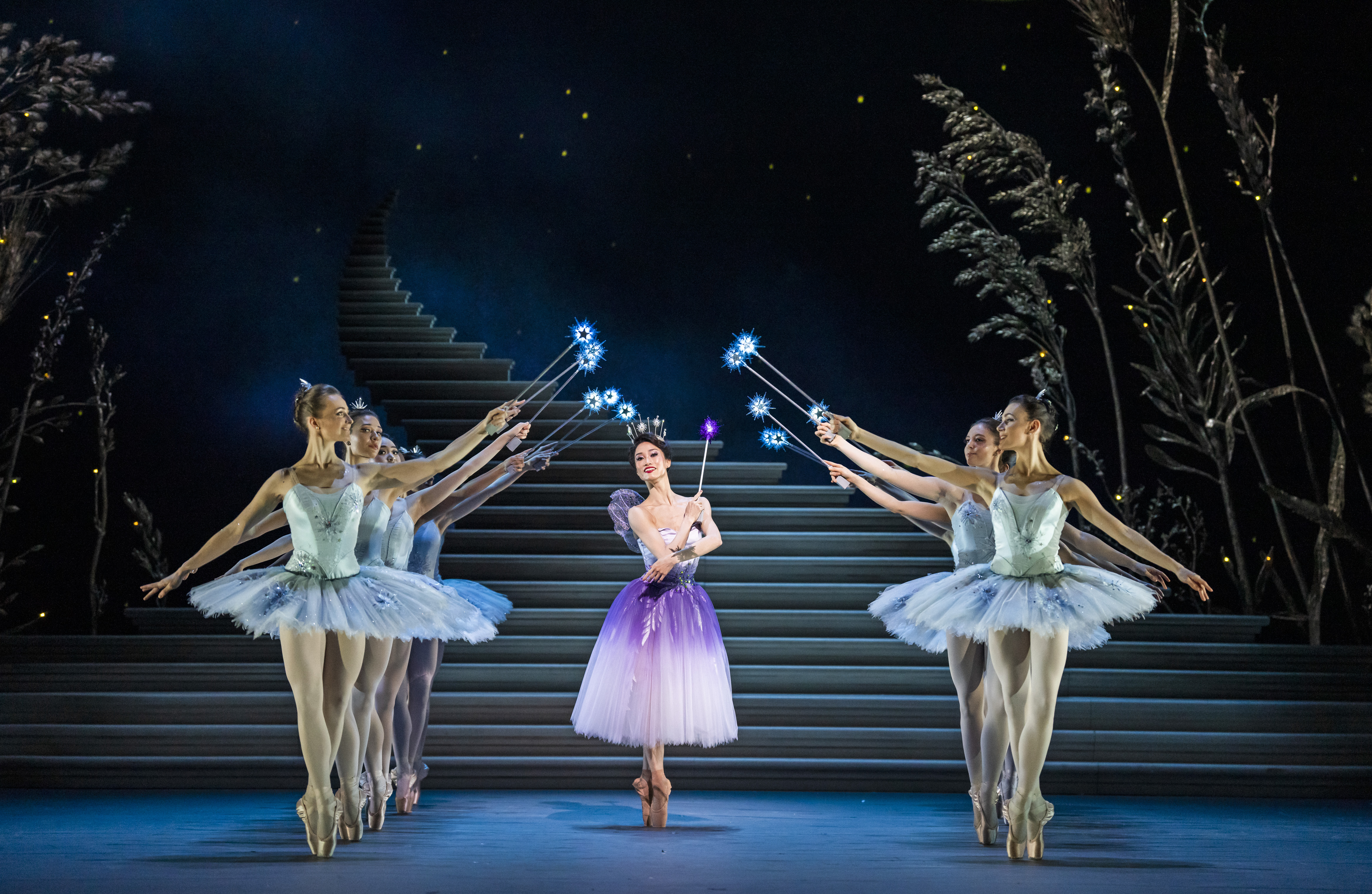 A ballet dancer in a white and purple dress, surrounded by eight dancers in light blue tutus, all holding wands with sparkling stars. The backdrop includes a staircase and a dark sky with subtle lighting effects, creating a magical atmosphere.