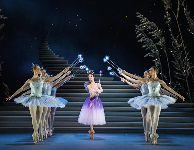 A ballet dancer in a white and purple dress, surrounded by eight dancers in light blue tutus, all holding wands with sparkling stars. The backdrop includes a staircase and a dark sky with subtle lighting effects, creating a magical atmosphere.