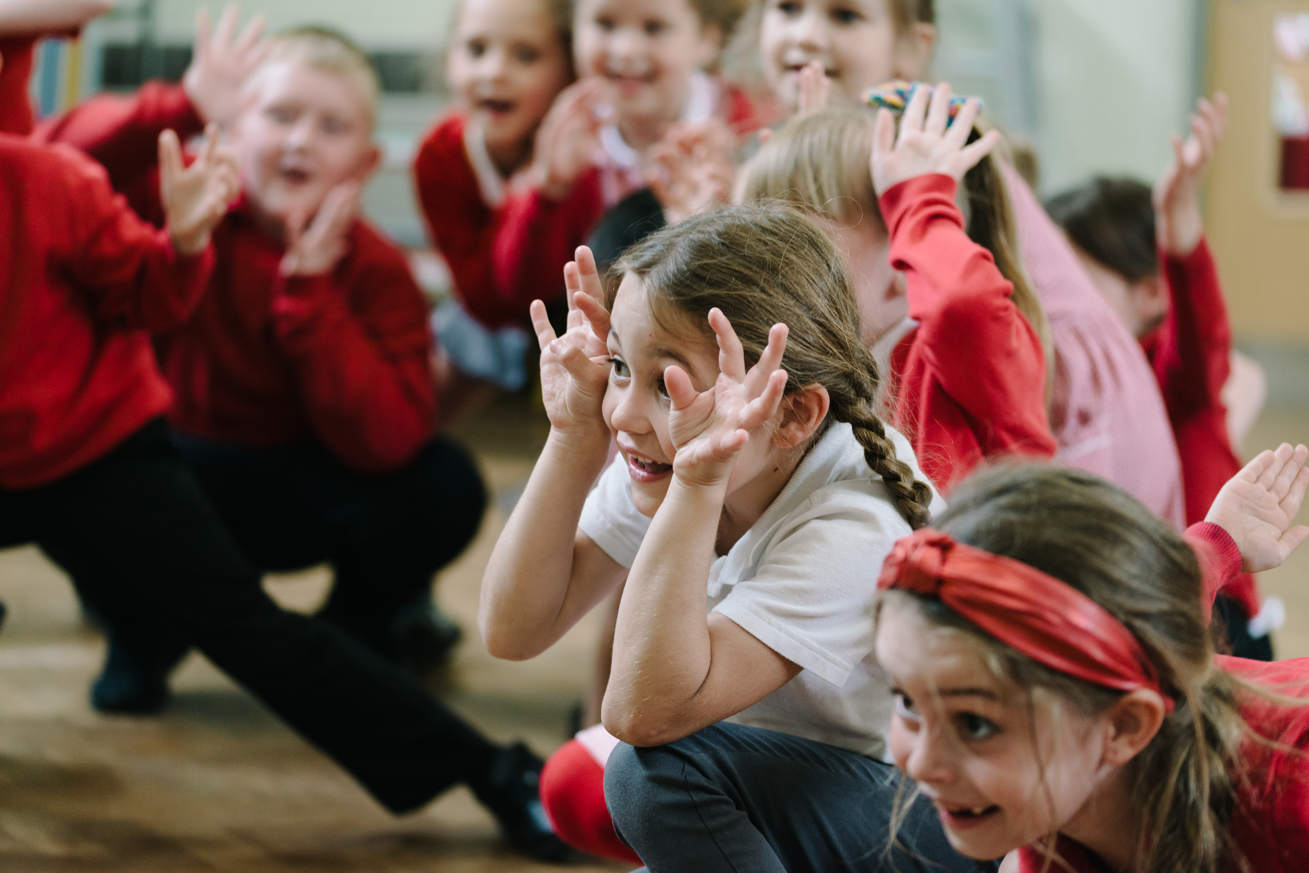 Girl in school uniform crouches in the foreground. Grinning and using her hands to create a funny expression. Other children in the background out of focus, are pulling similar funny faces. 