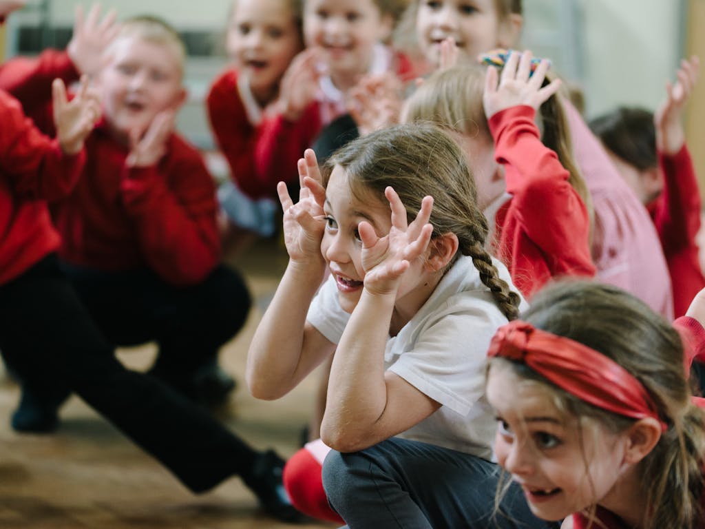 Girl in school uniform crouches in the foreground. Grinning and using her hands to create a funny expression. Other children in the background out of focus, are pulling similar funny faces.