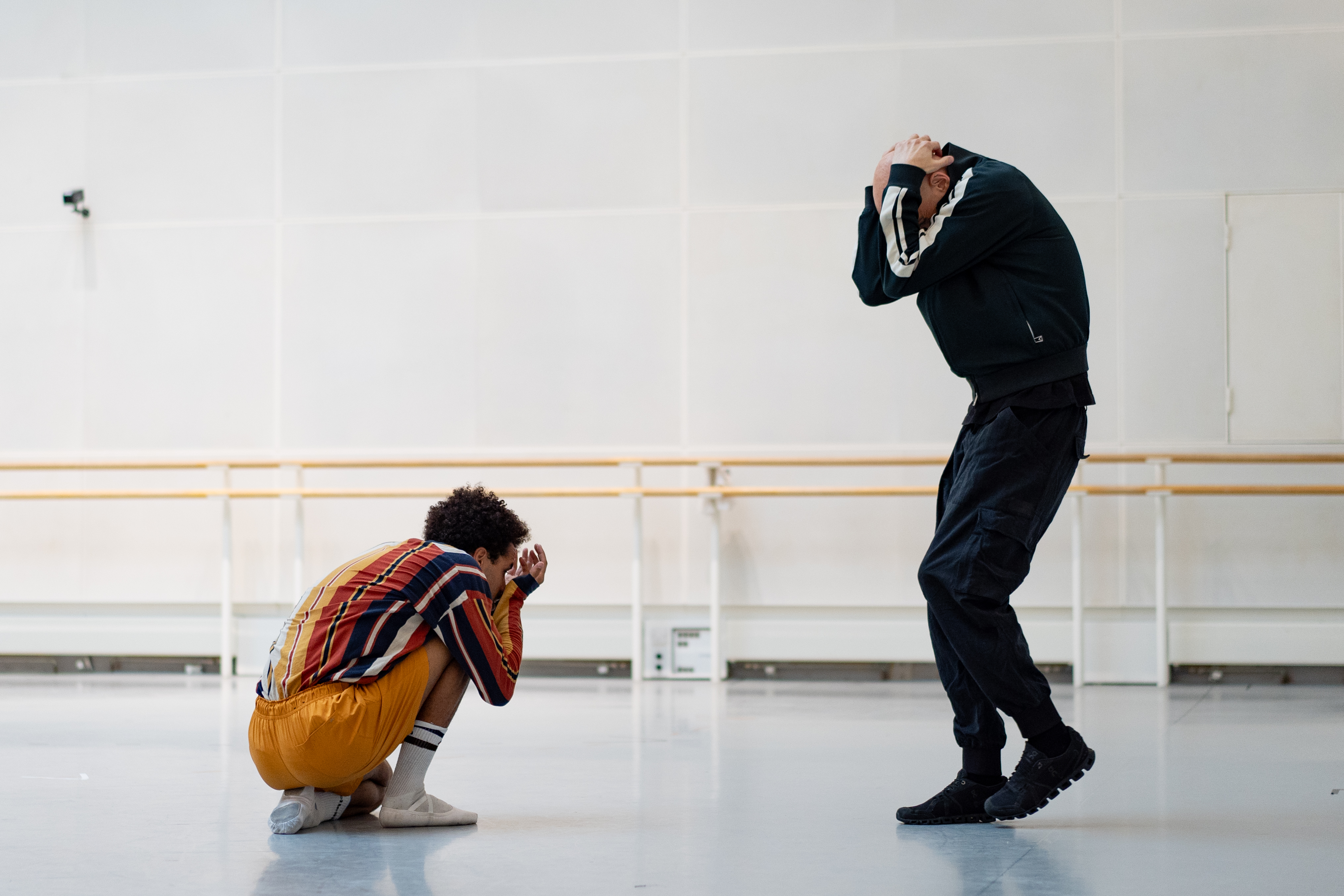 Two dancers rehearse in a white ballet studio. One is dressed in orange and crouches on the floor, the other is wearing a black tracksuit. Both have their heads in their hands. 