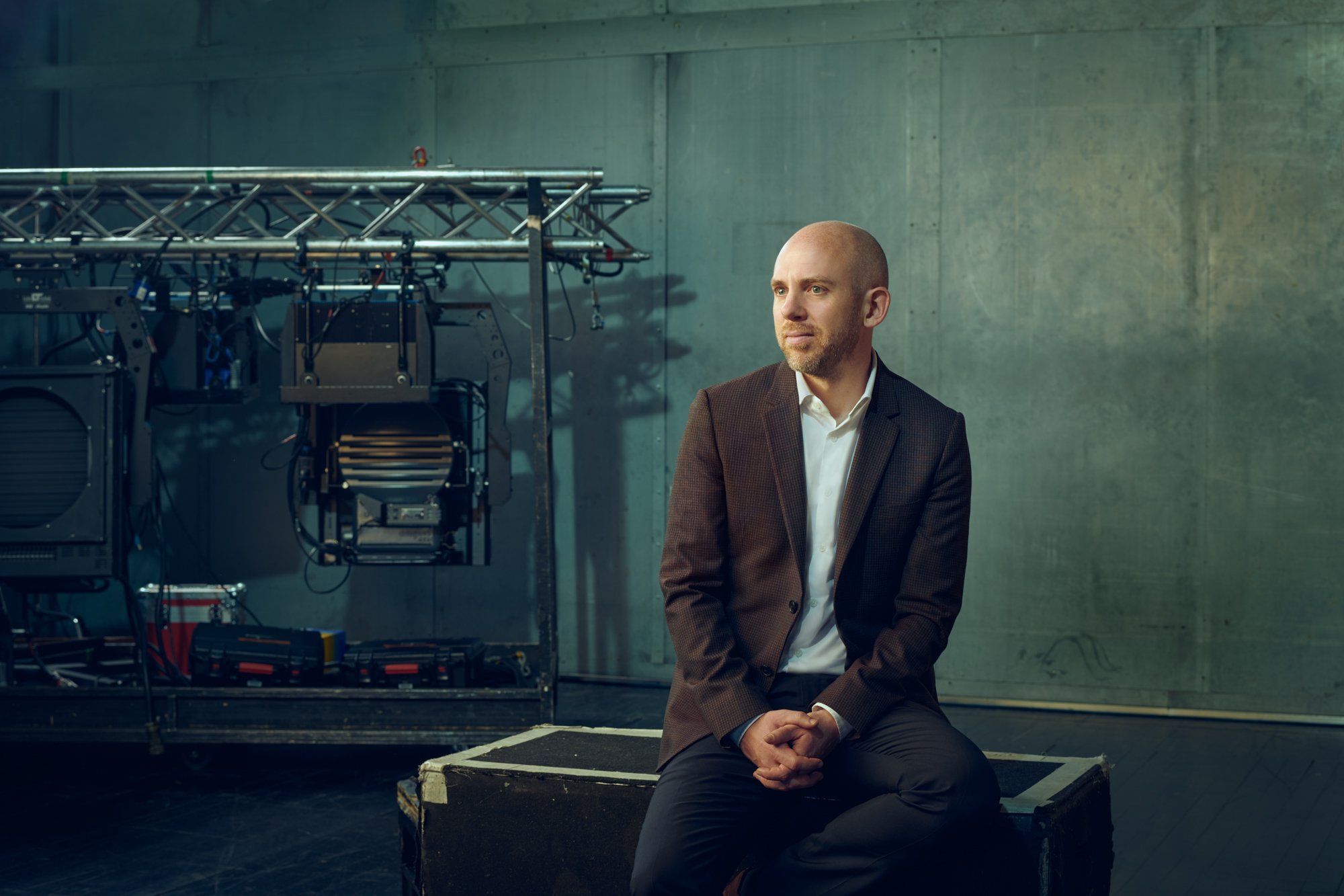 Director of the Royal Opera, Oliver Mears, is wearing a brown blazer and sits on a wooden crate, looking to the side. Behind him, there are professional lighting and audio equipment in front of muted green walls.