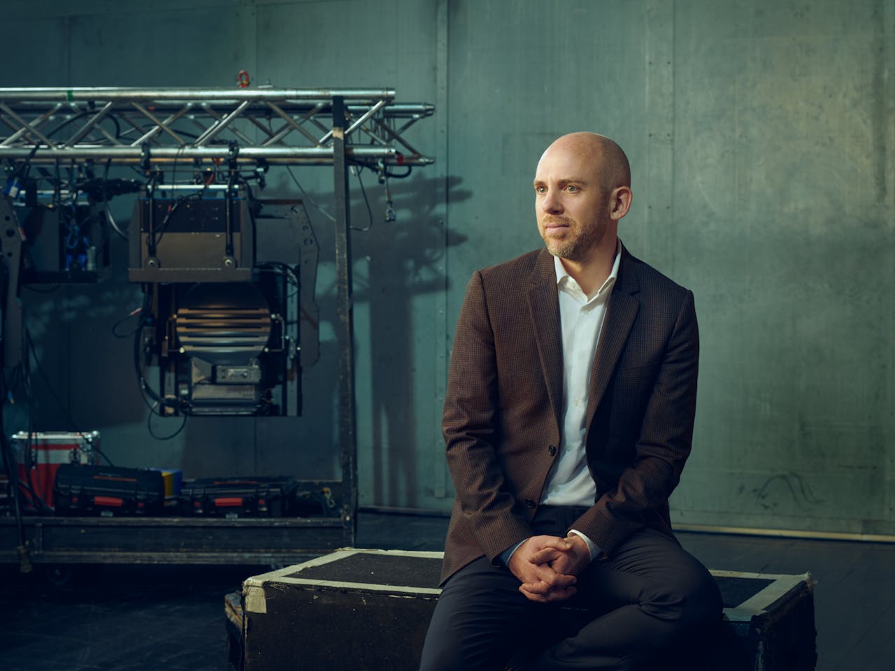 Oliver Mears, wearing a brown blazer sits on a wooden crate, looking to the side. Behind him, there are professional lighting and audio equipment in front of muted green walls.