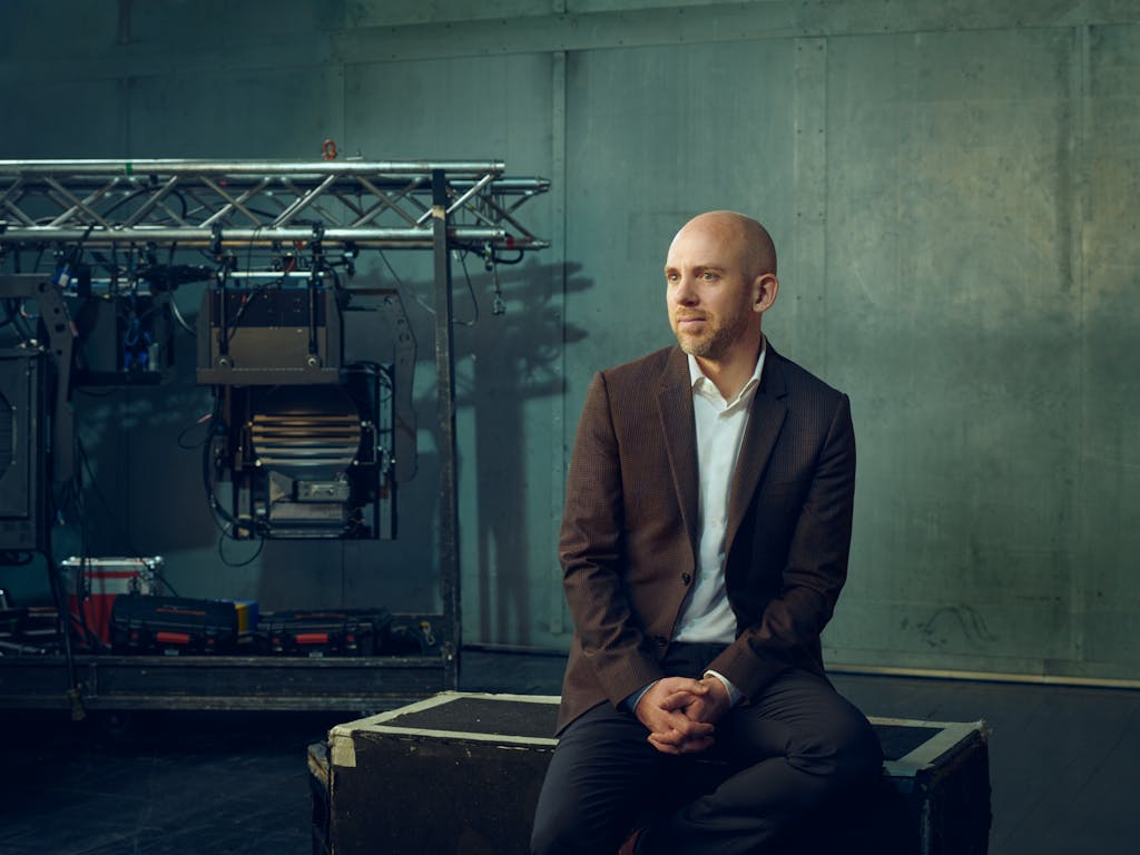 Director of the Royal Opera, Oliver Mears, is wearing a brown blazer and sits on a wooden crate, looking to the side. Behind him, there are professional lighting and audio equipment in front of muted green walls.