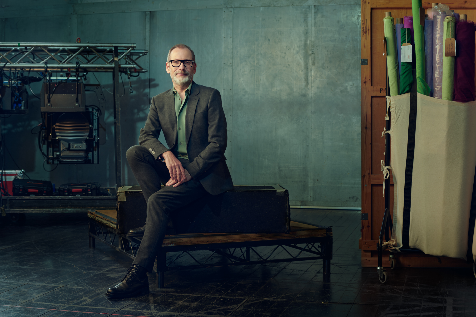 Kevin O'Hare, wearing glasses, sits on a bench in a studio, dressed in a grey suit and green shirt. Behind him, equipment, including lights and rolls of colourful fabric, are leant against muted green walls.