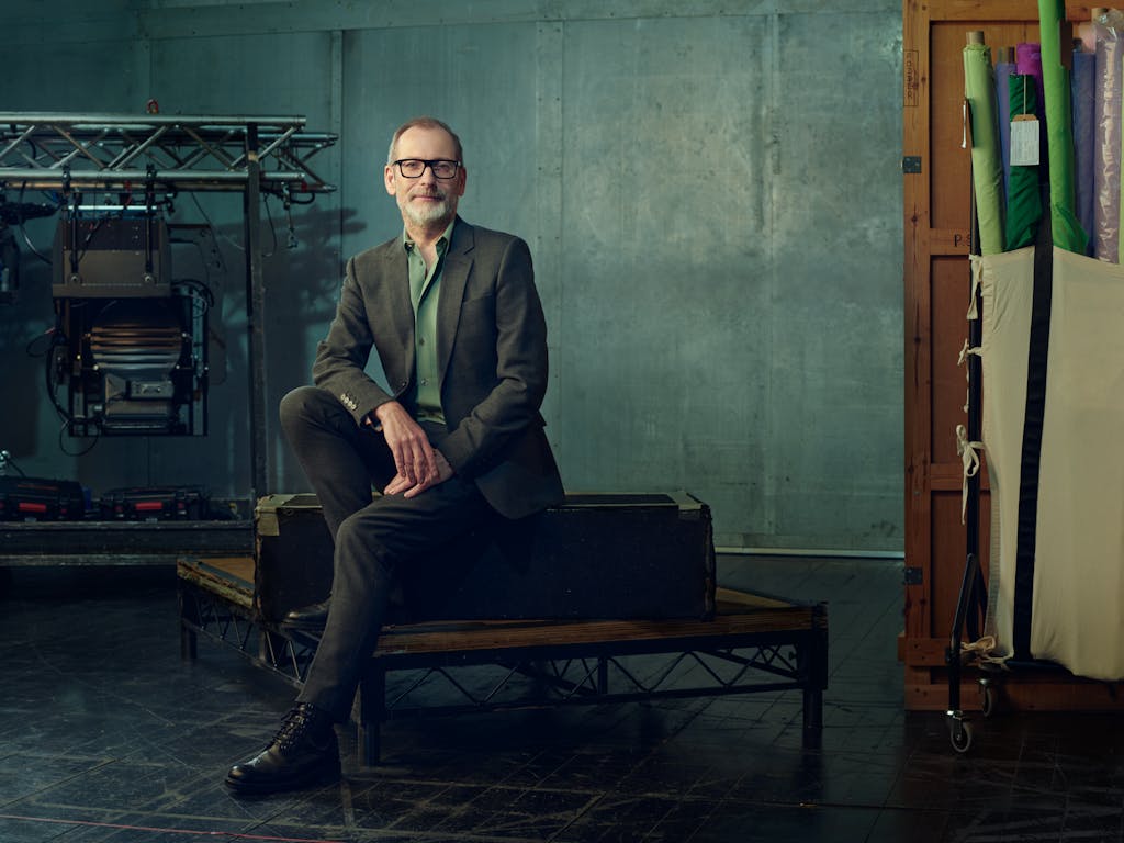 Kevin O'Hare, wearing glasses, sits on a bench in a studio, dressed in a grey suit and green shirt. Behind him, equipment, including lights and rolls of colourful fabric, are leant against muted green walls.