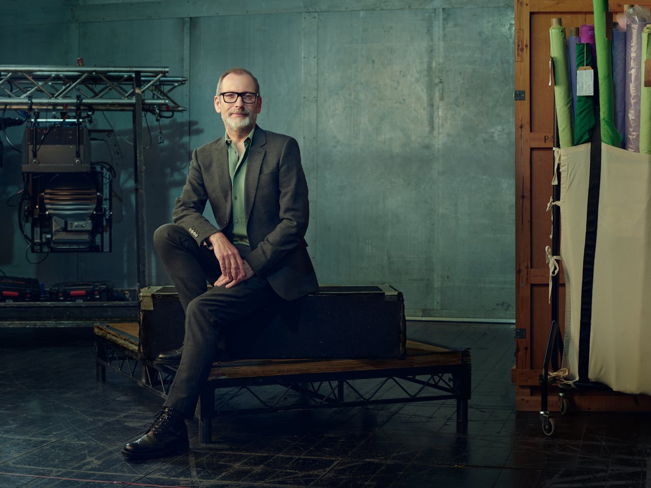Kevin O'Hare, wearing glasses, sits on a bench in a studio, dressed in a grey suit and green shirt. Behind him, equipment, including lights and rolls of colourful fabric, are leant against muted green walls.