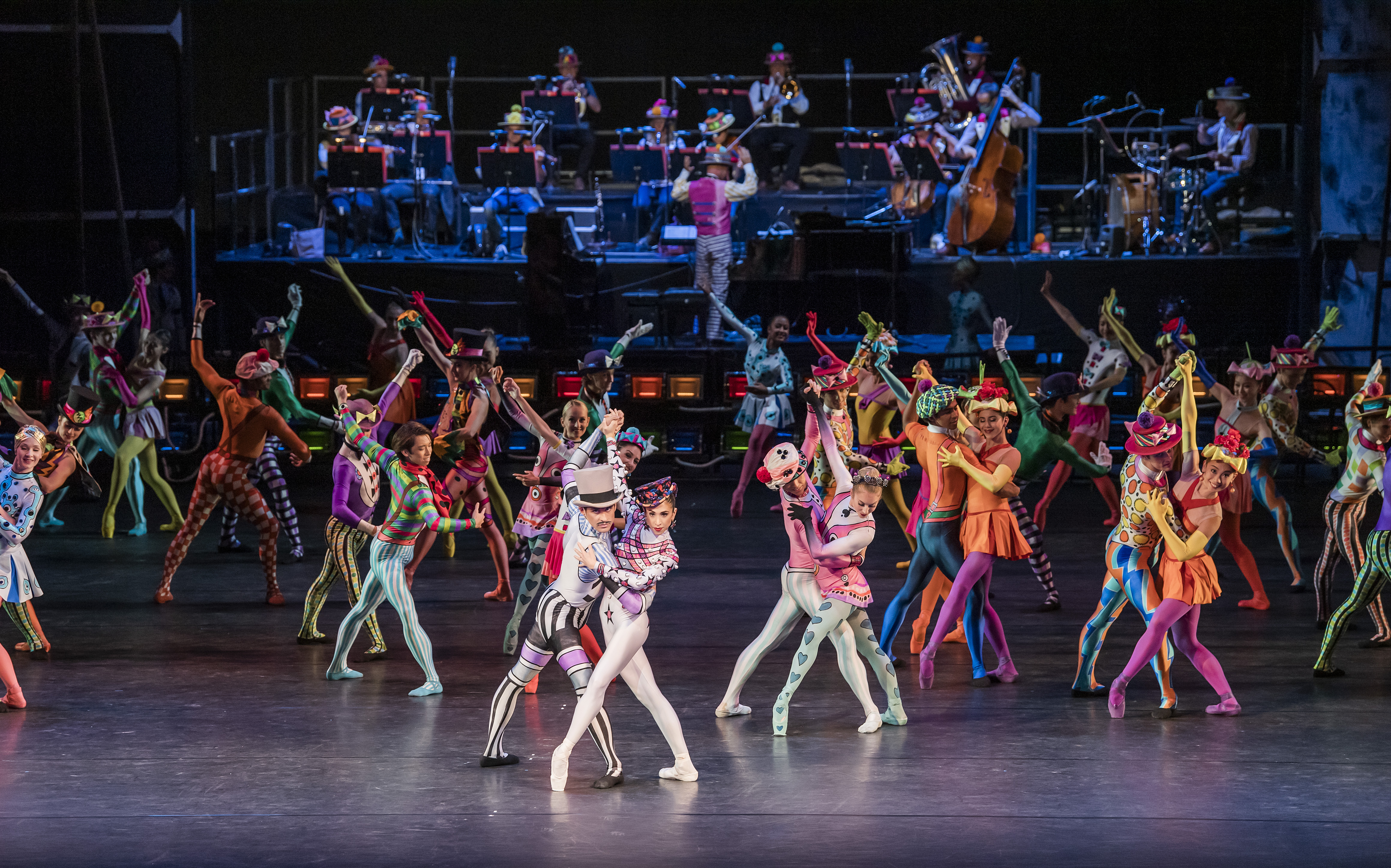 The Royal Ballet perform Elite Syncopations. Dancers on stage in colourful costumes, including polka dots and stripes, pose in pairs. A band performs on a raised platform behind them.