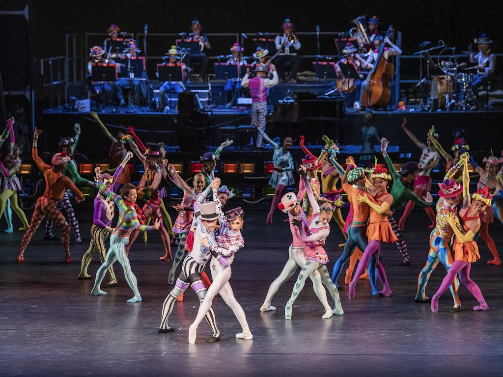 The Royal Ballet perform Elite Syncopations. Dancers on stage in colourful costumes, including polka dots and stripes, pose in pairs. A band performs on a raised platform behind them.