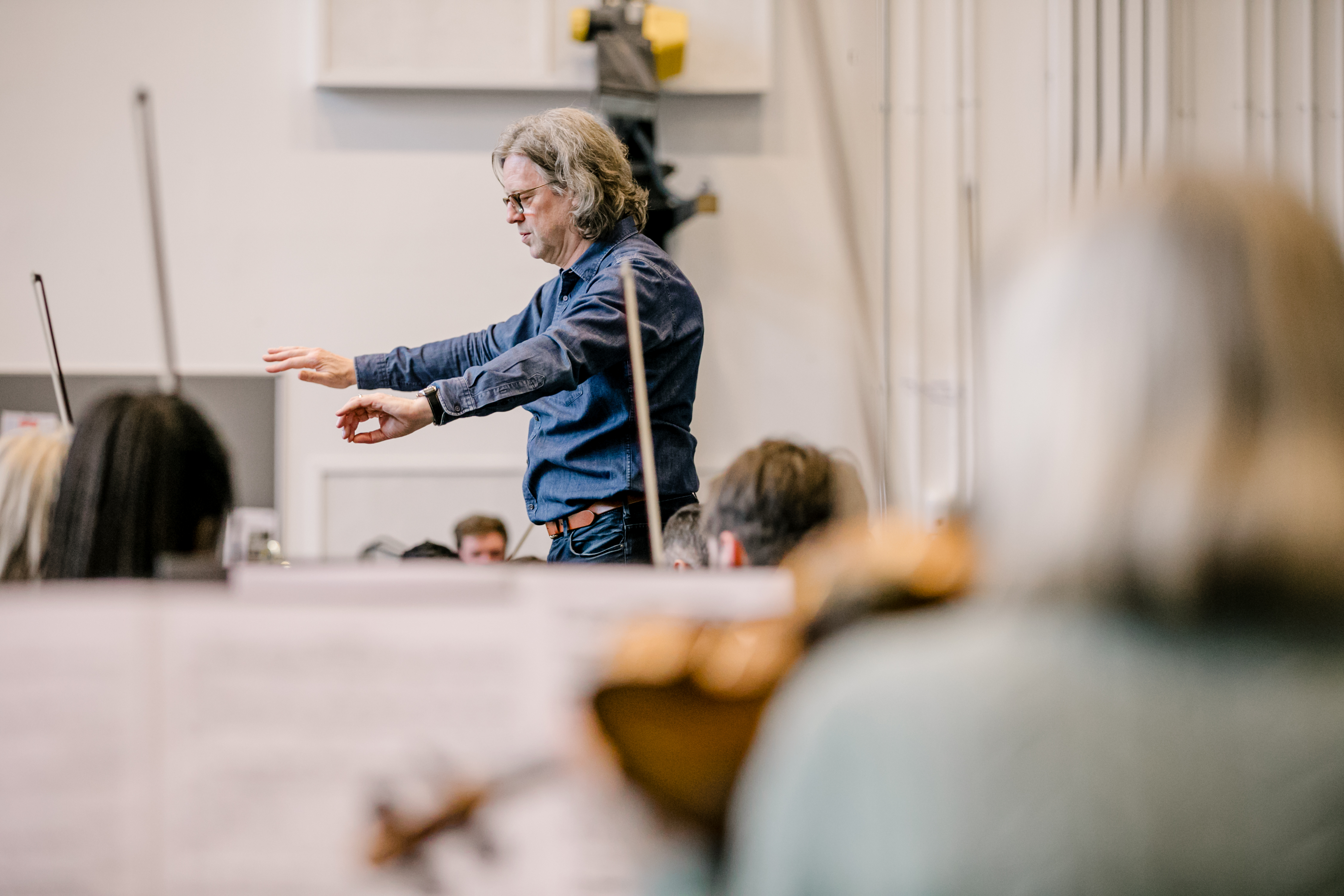 A conductor, Koen Kessels, leads an orchestra,gesturing with both hands, while string instruments are blurred in the foreground. The setting is a softly lit rehearsal space.