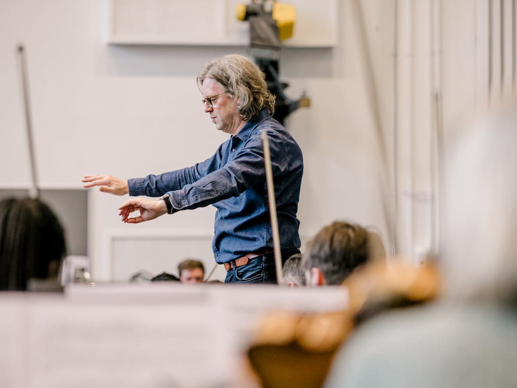 A conductor, Koen Kessels, leads an orchestra,gesturing with both hands, while string instruments are blurred in the foreground. The setting is a softly lit rehearsal space.