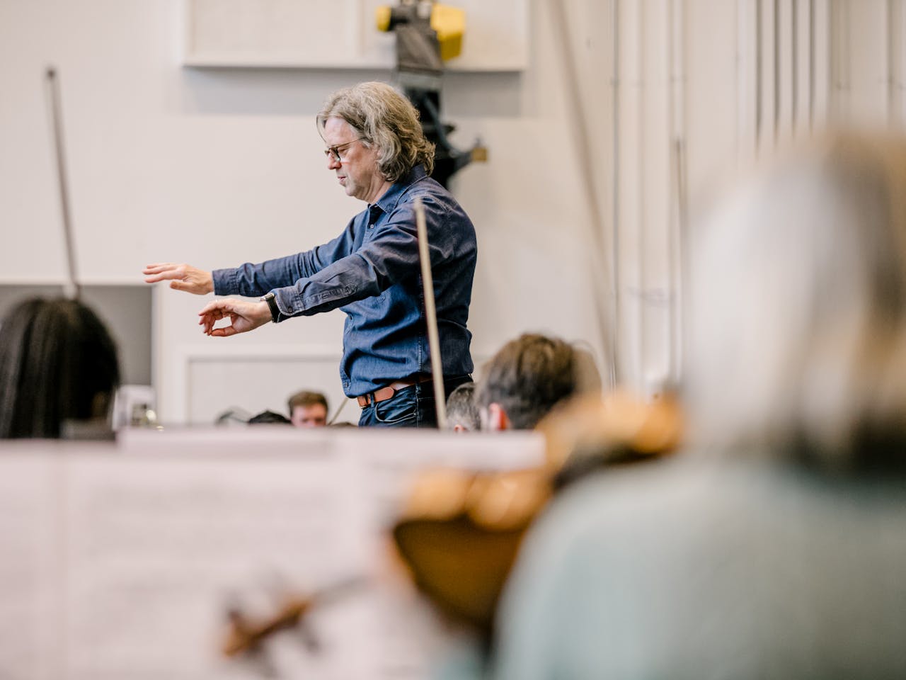 A conductor, Koen Kessels, leads an orchestra,gesturing with both hands, while string instruments are blurred in the foreground. The setting is a softly lit rehearsal space.