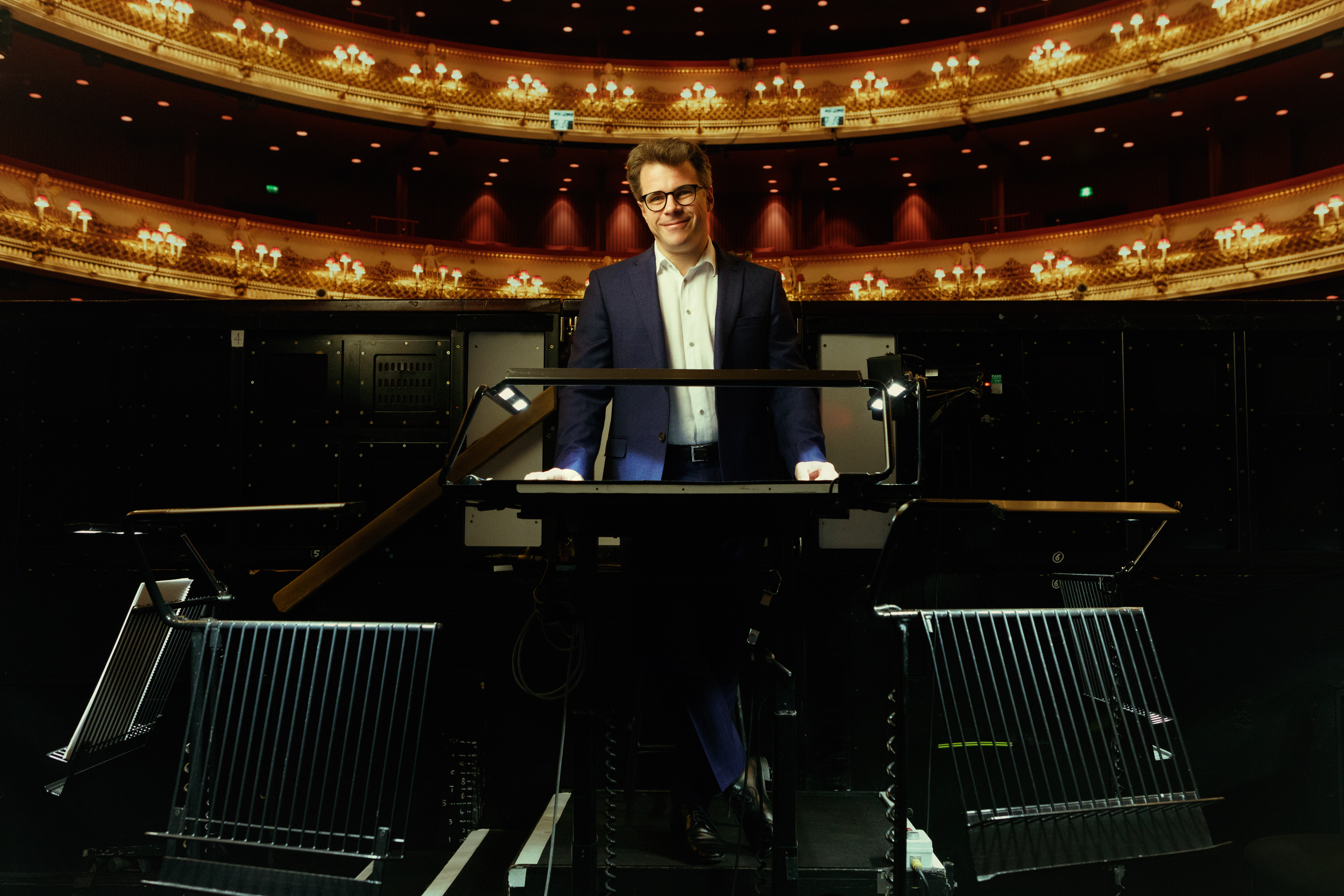 Jakub Hrůša, wearing a dark suit and glasses, stands behind a conductor's podium in the orchestra pit at the Royal Opera House. An ornate balcony and chandeliers are visible in the background as he smiles at the camera.