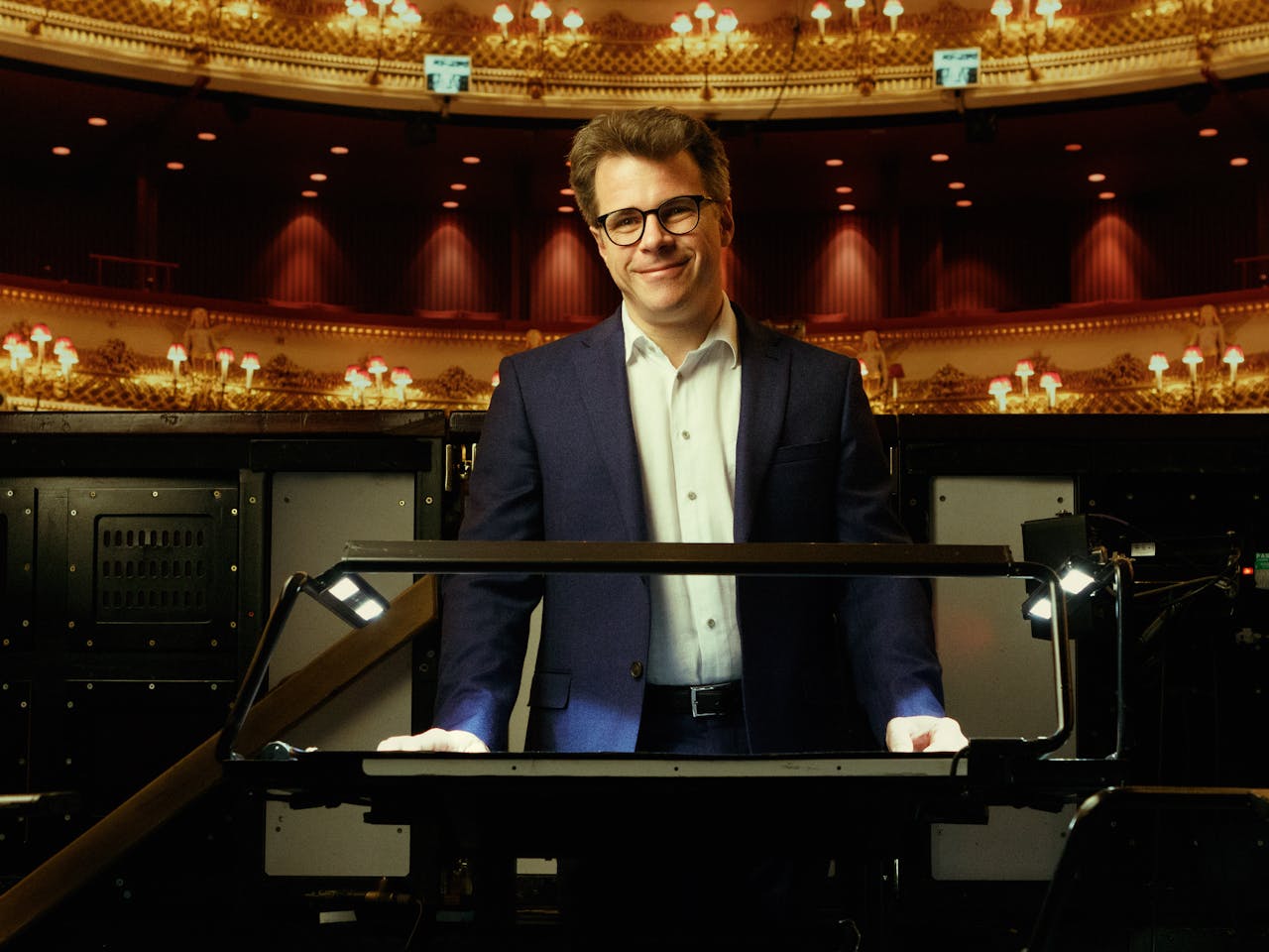 Jakub Hrůša, wearing a dark suit and glasses, stands behind a conductor's podium in the orchestra pit at the Royal Opera House. An ornate balcony and chandeliers are visible in the background as he smiles at the camera.
